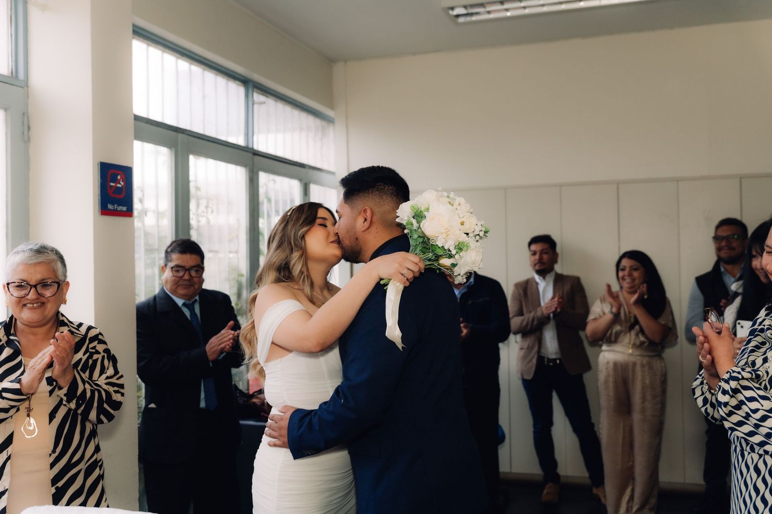 Newlyweds share first kiss as wedding guests applaud at civil ceremony.
