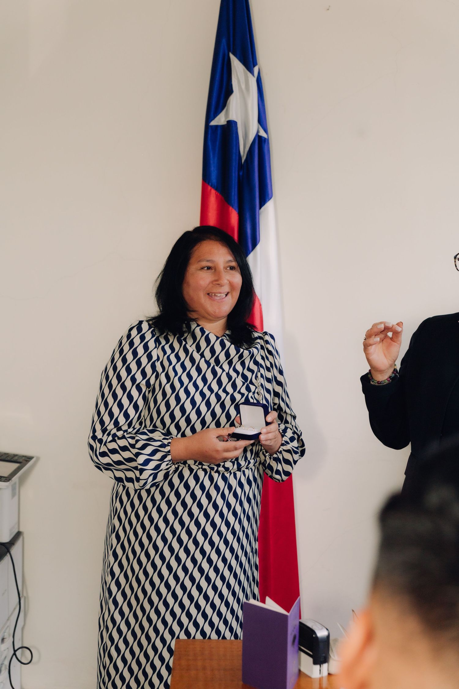 Woman in patterned dress holding ring box beside Chilean flag at wedding.