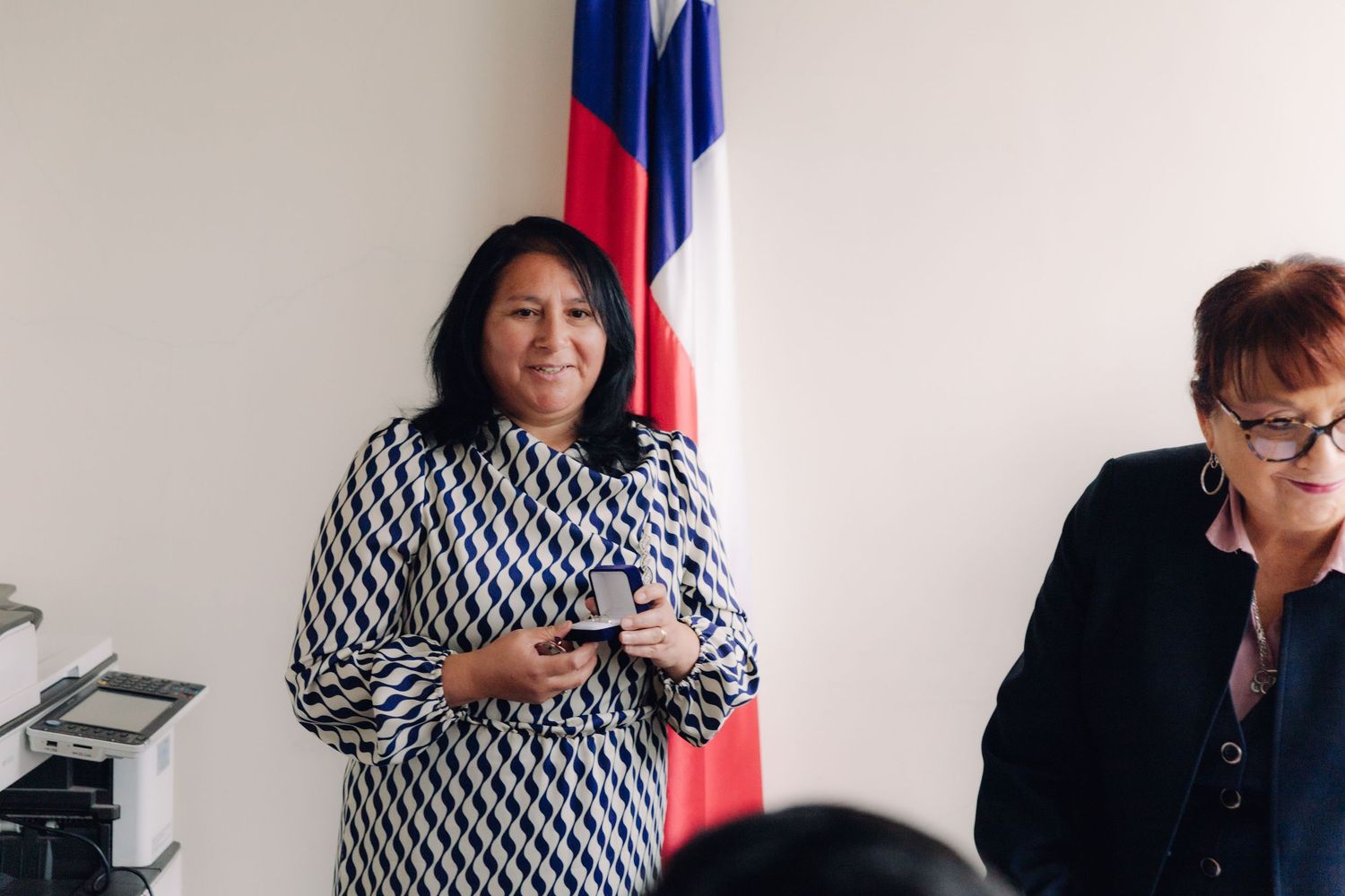 Woman in geometric dress smiling while holding ring box near Chilean flag.