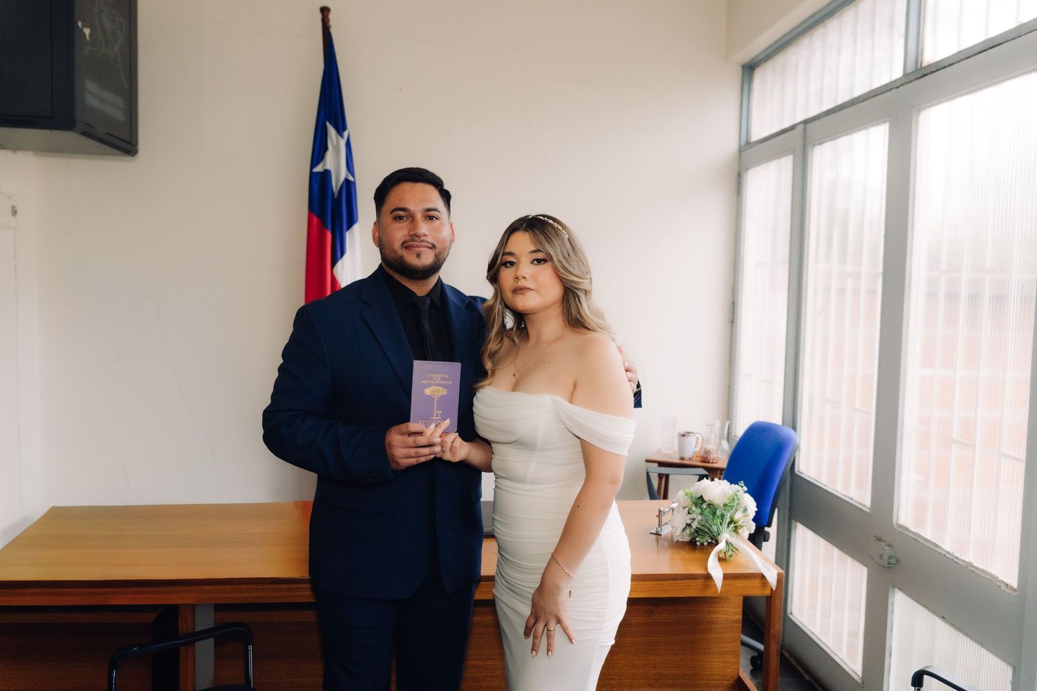 Newlywed couple poses with marriage certificate and bridal bouquet at Chilean civil ceremony.