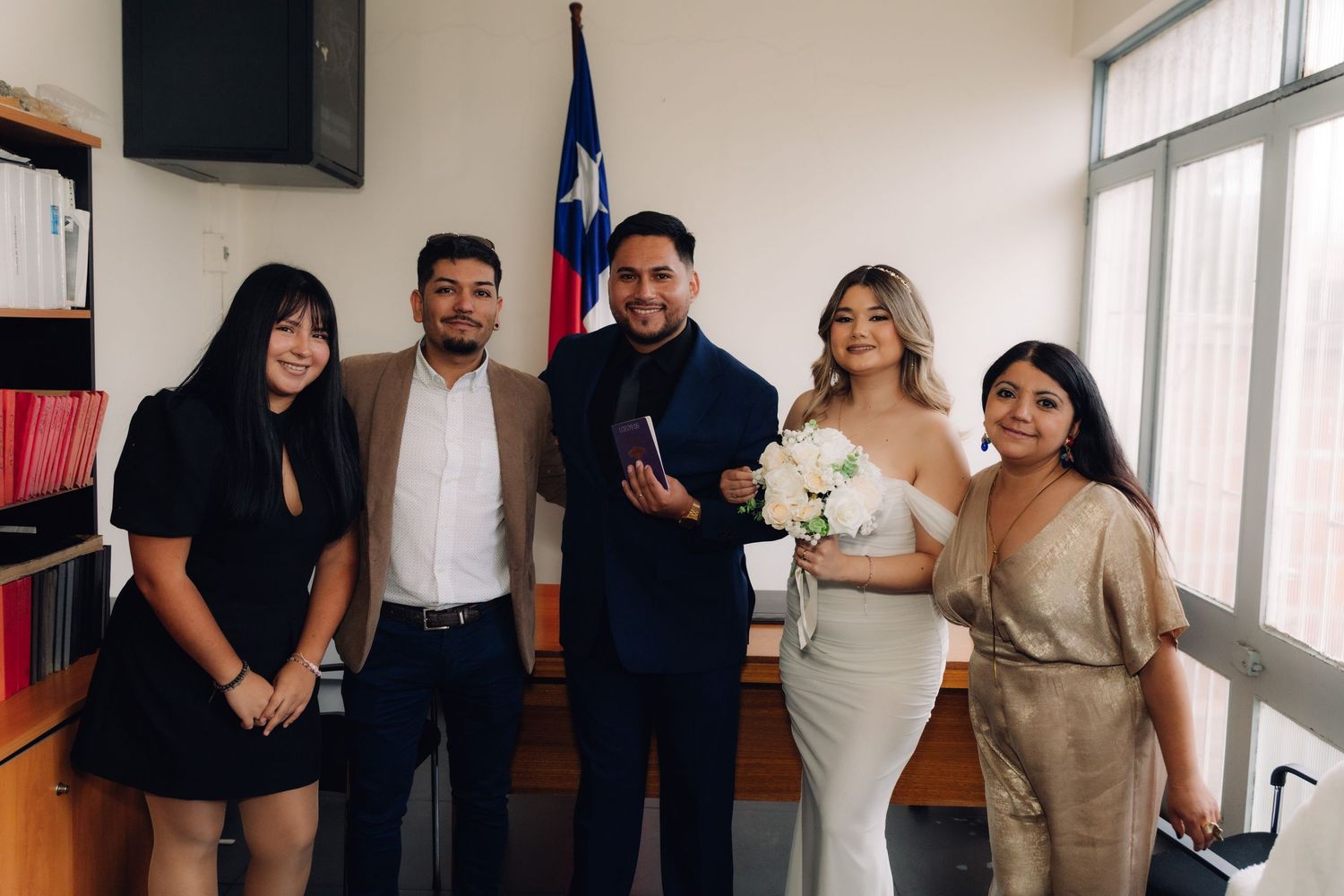 Couple in wedding attire poses with three guests in front of Chilean flag during civil ceremony.