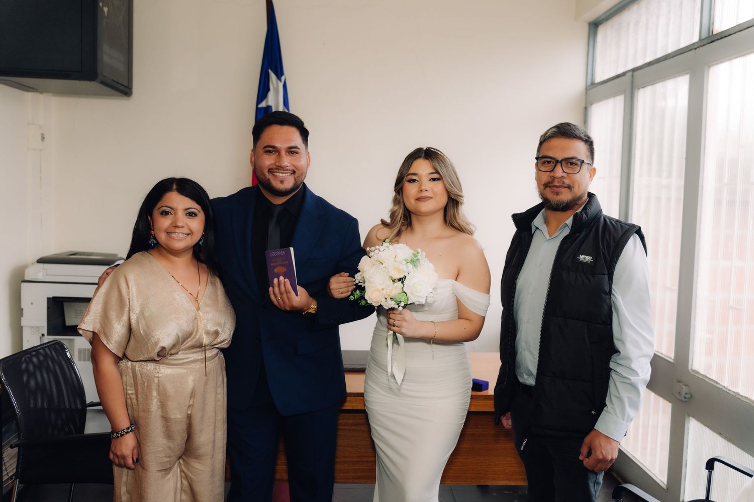 Bride in white gown and groom in navy suit pose with two guests at civil wedding ceremony.