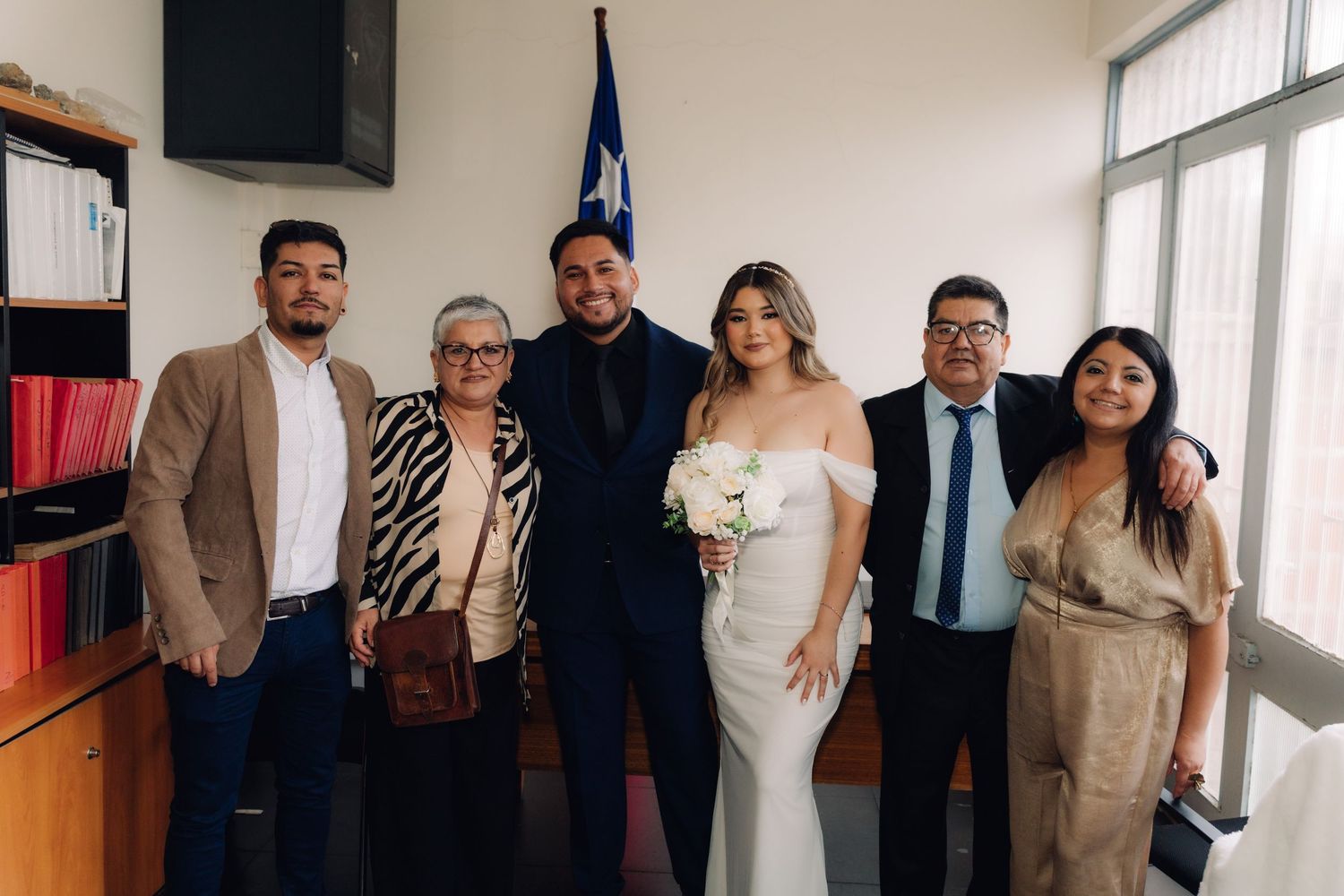 Wedding couple poses with four family members in front of Chilean flag during civil ceremony.