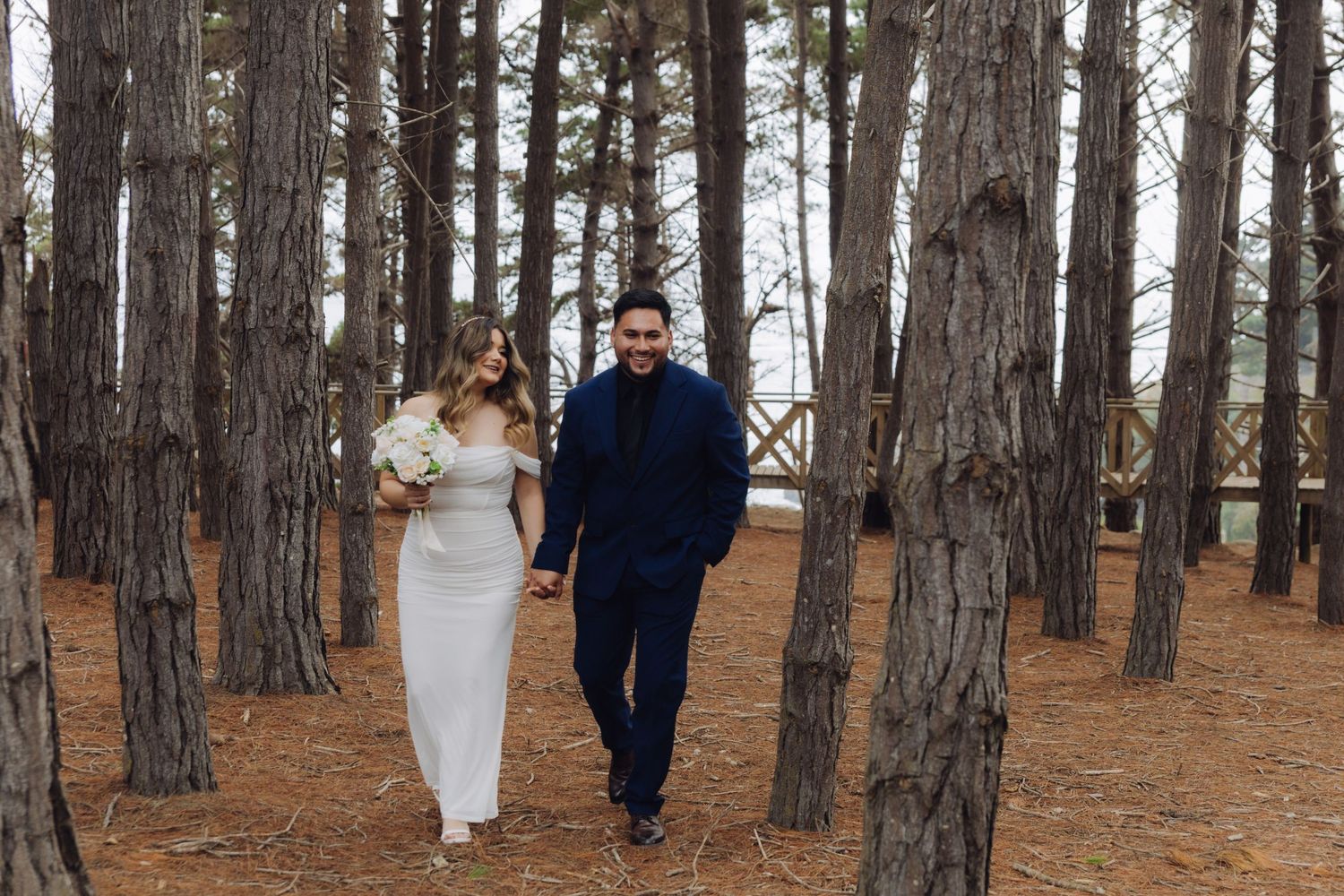 Newlyweds walk hand in hand through a scenic pine forest in wedding attire.