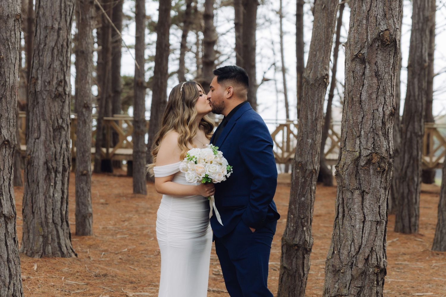 Bride and groom share a romantic kiss among tall pine trees in a forest wedding photo.