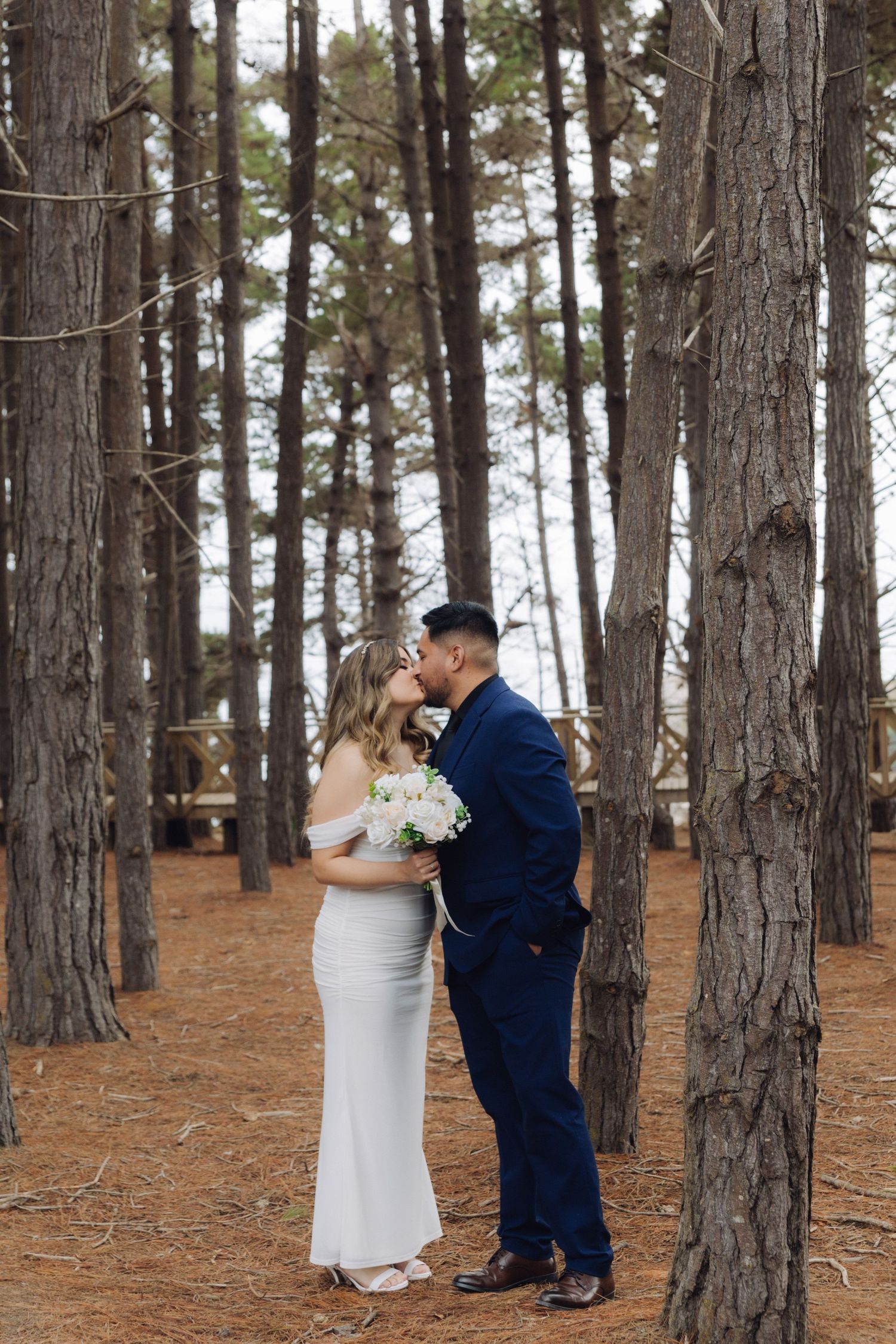 Bride and groom share a tender kiss surrounded by tall pine trees in forest setting.