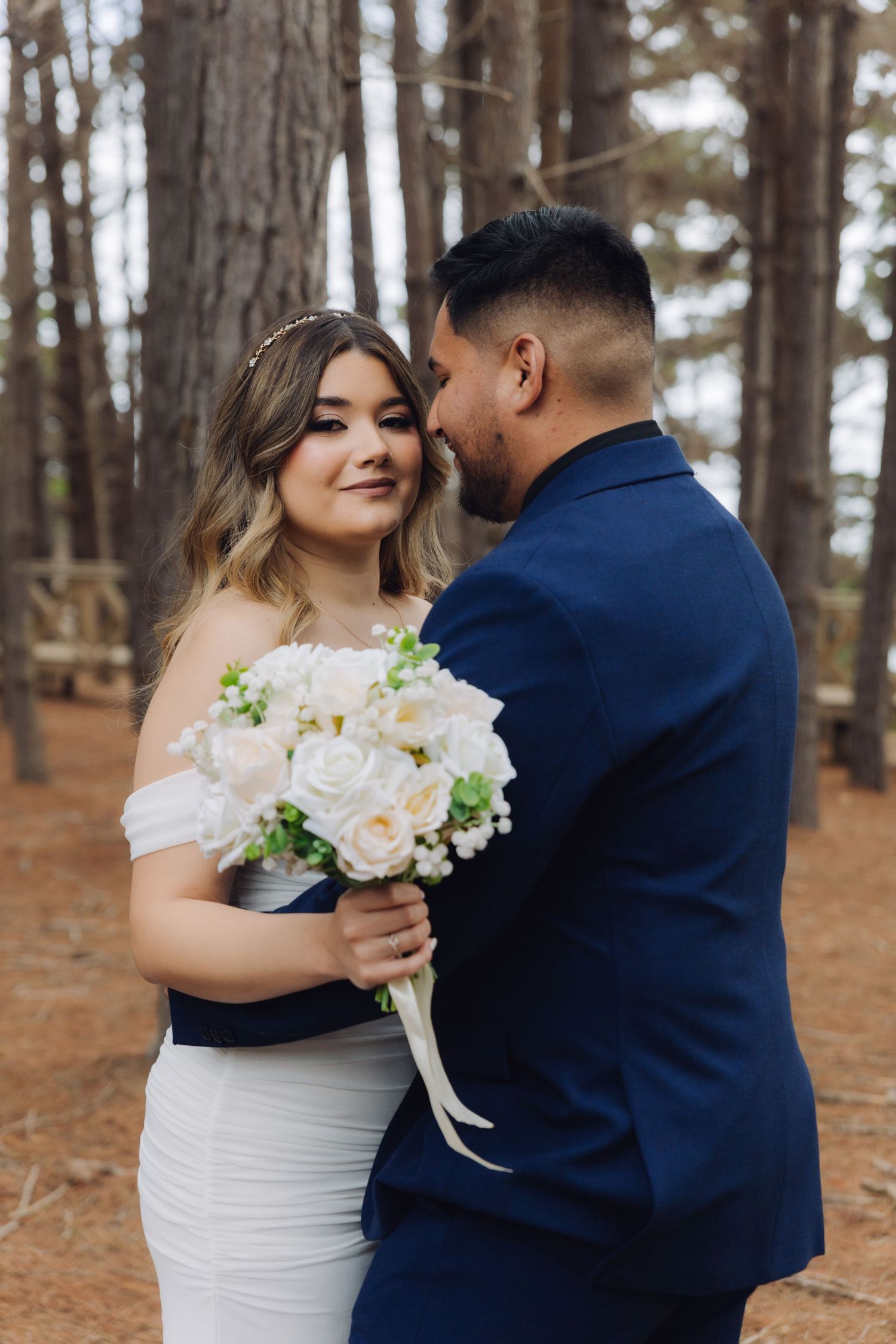 Bride smiles holding white rose bouquet as groom leans close in a pine forest setting.