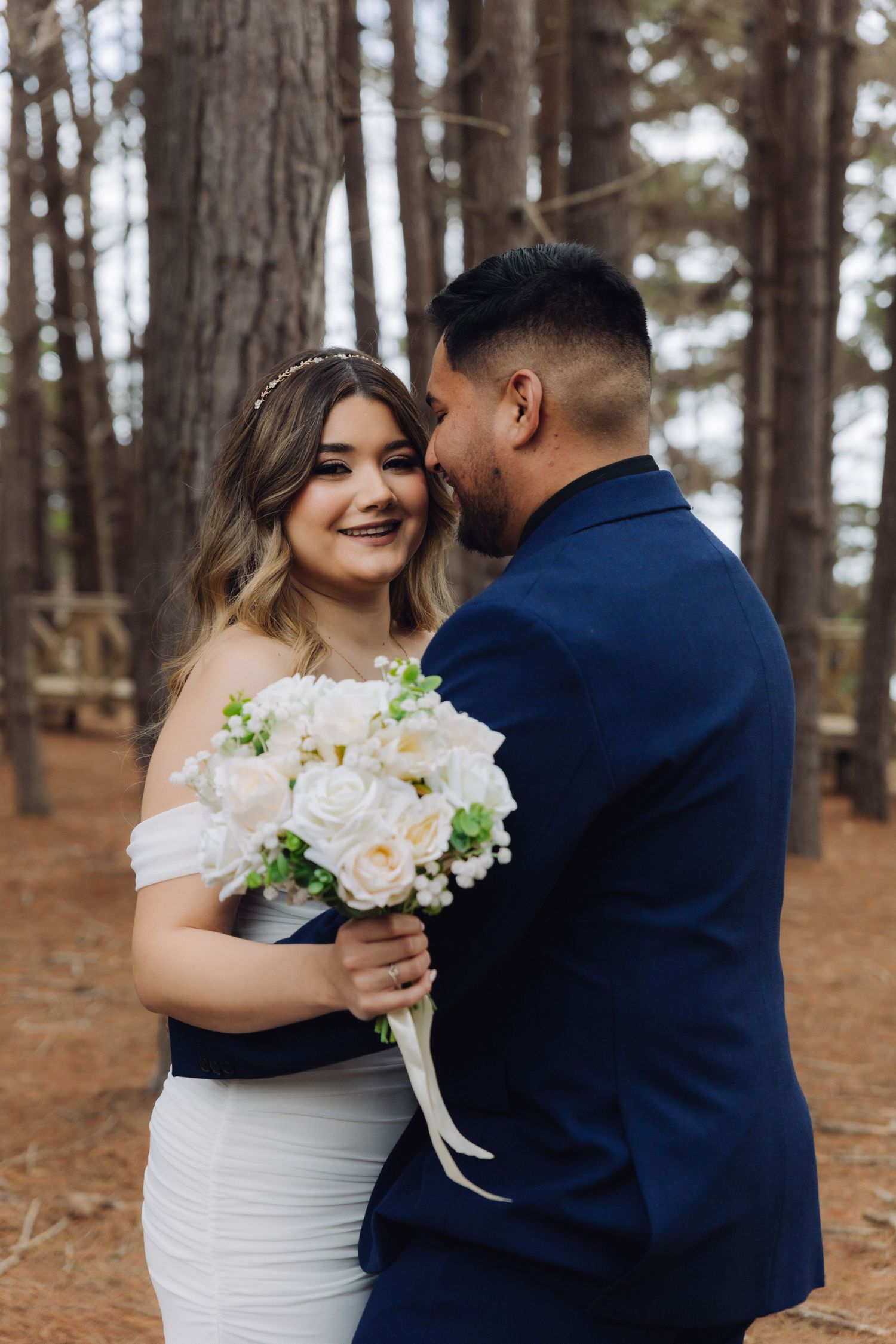 Smiling bride in off-shoulder gown holds white bouquet while groom whispers in her ear outdoors.