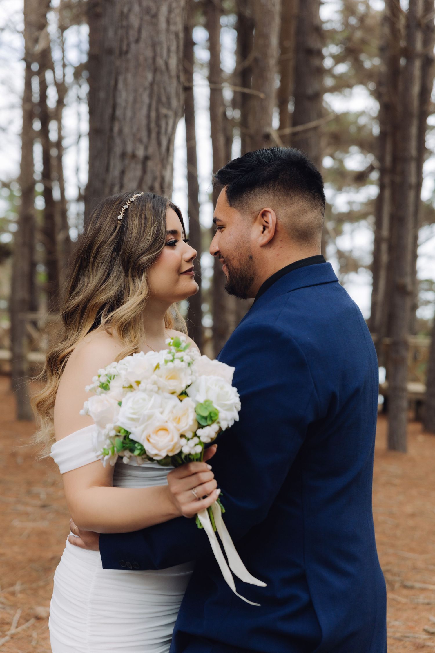 Bride and groom gaze lovingly at each other holding white floral bouquet in pine forest.