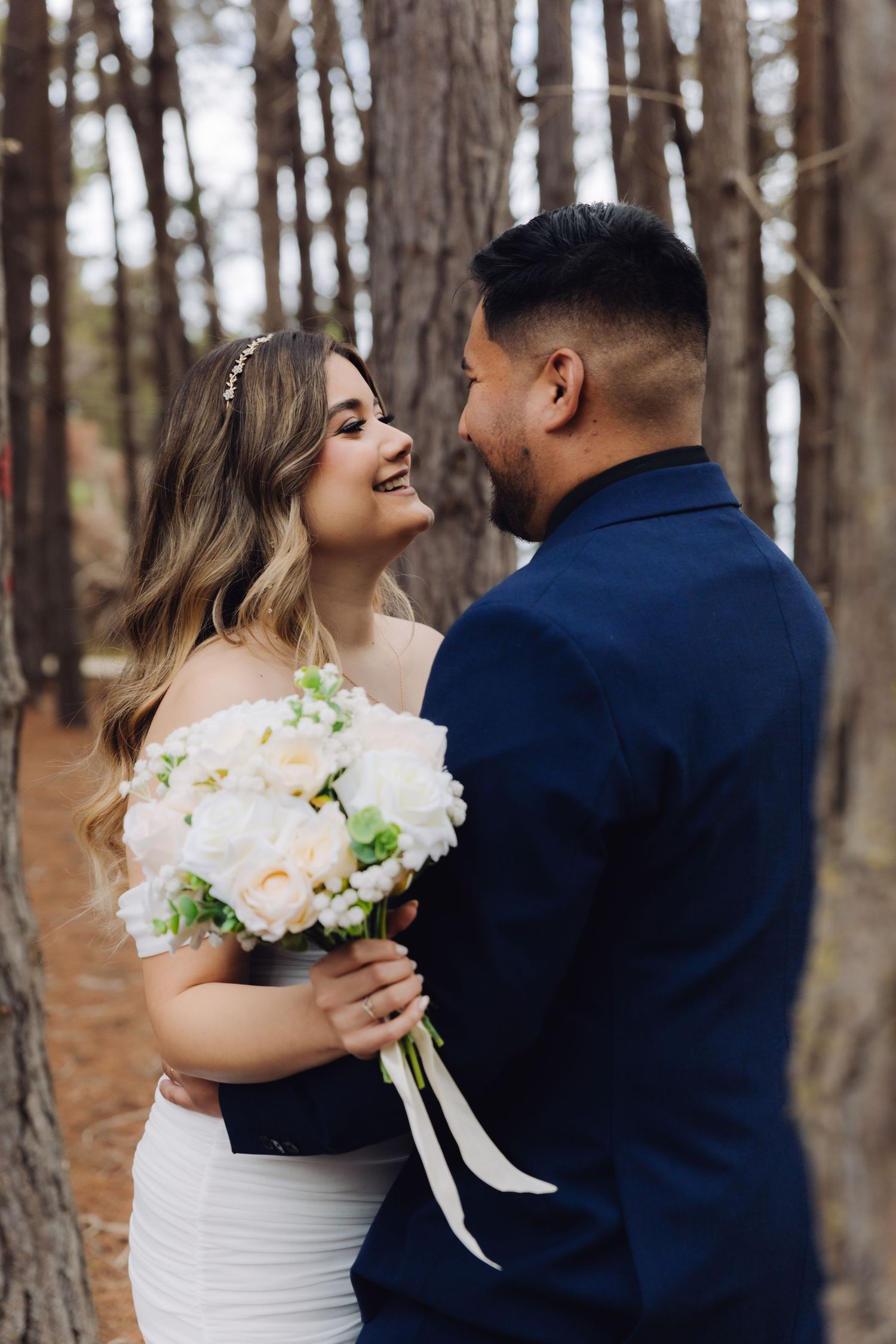 Bride and groom share a romantic moment in a pine forest, bride holding white peony bouquet in off-shoulder gown.