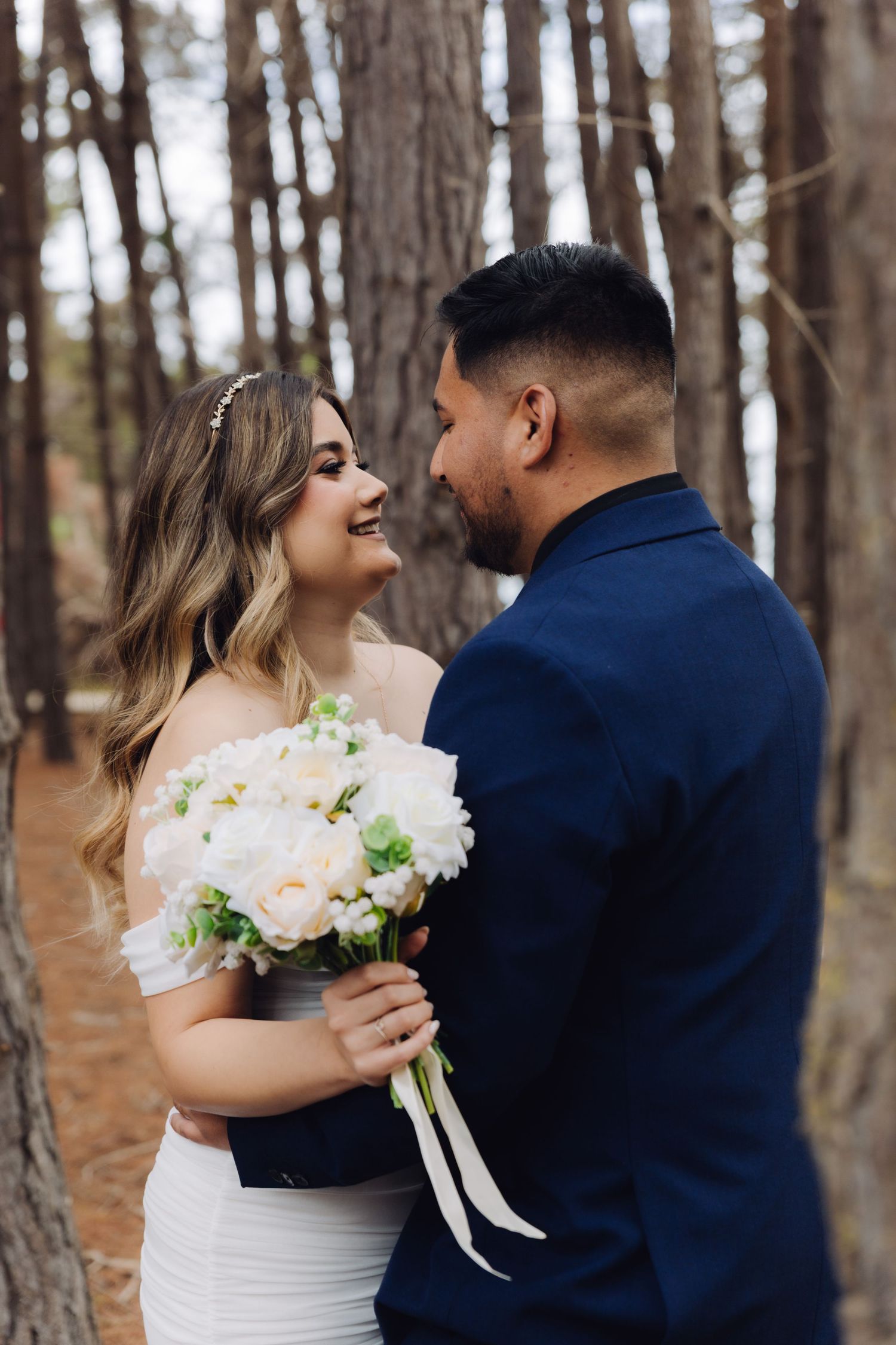 Bride laughs joyfully as groom gazes at her while she holds a white bouquet in forest.
