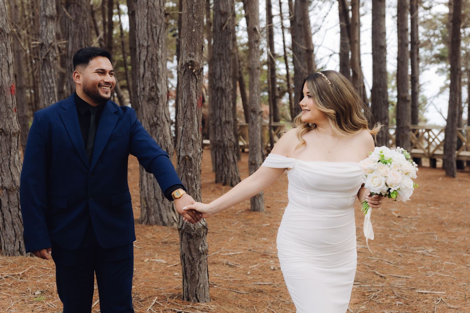 Wedding couple holds hands walking through pine forest, groom in navy suit and bride in white off-shoulder dress.