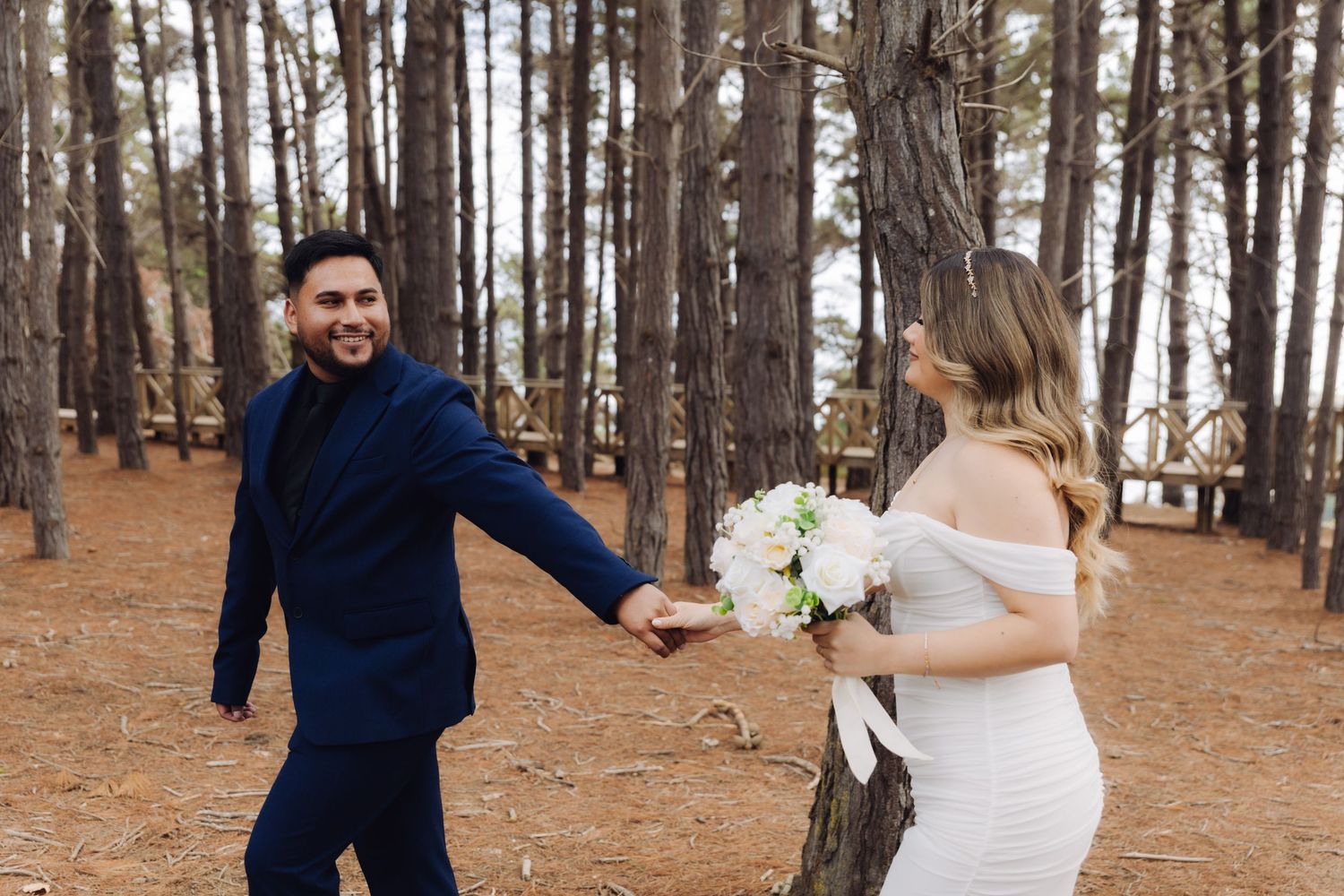 Smiling groom in navy suit leads bride through sunlit pine forest as she carries a white floral bouquet.