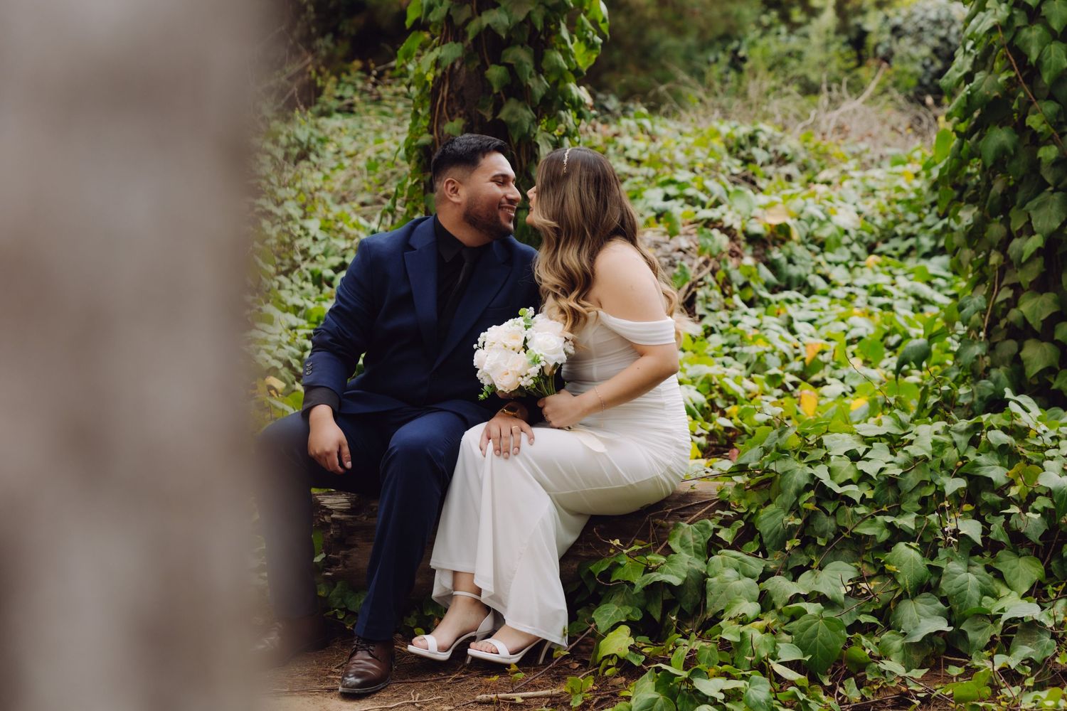 Bride and groom share a kiss seated on a stone bench surrounded by lush green ivy in a garden setting.