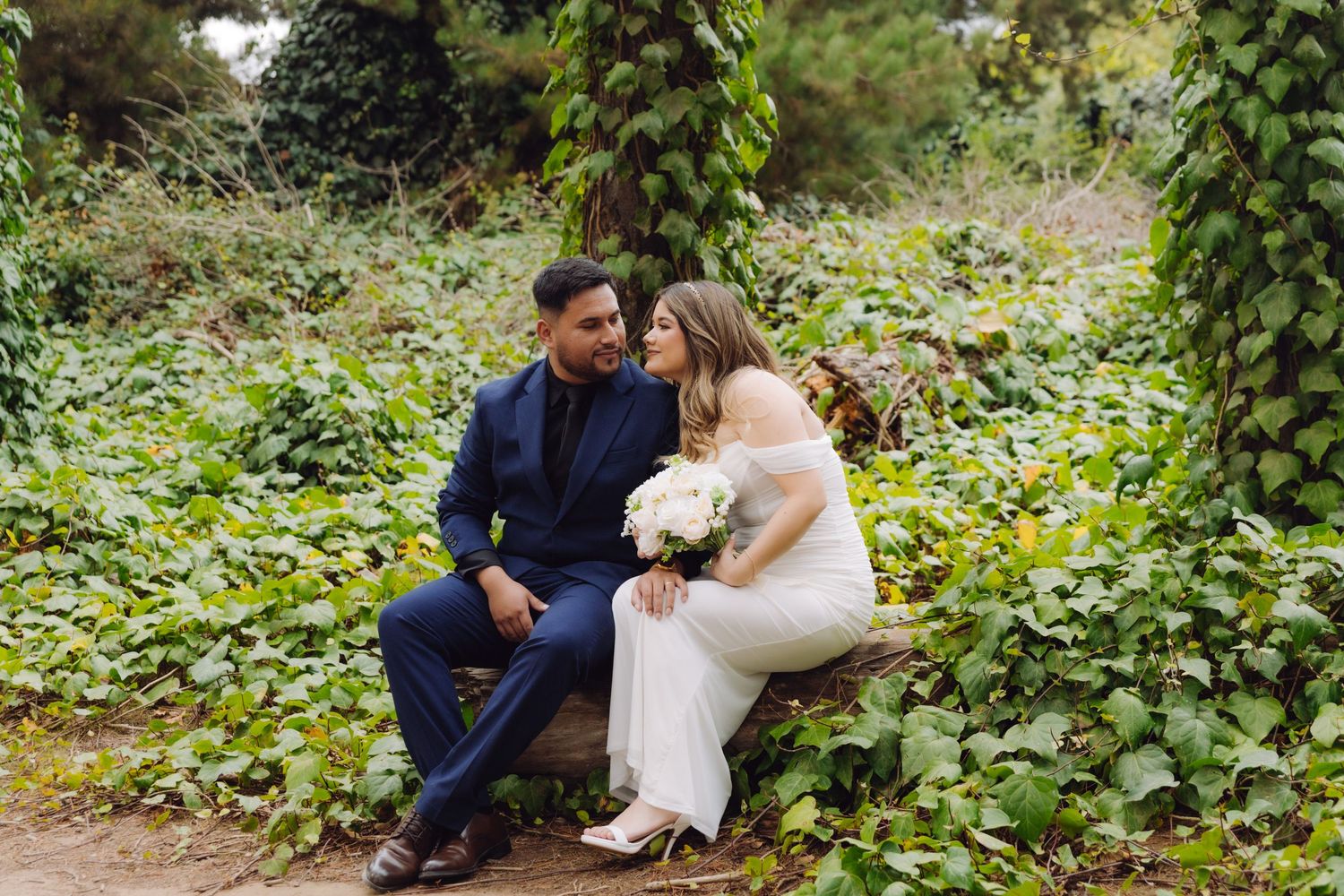 Elegant wedding couple sitting on a rustic bench gazing at each other amid vibrant green garden foliage.