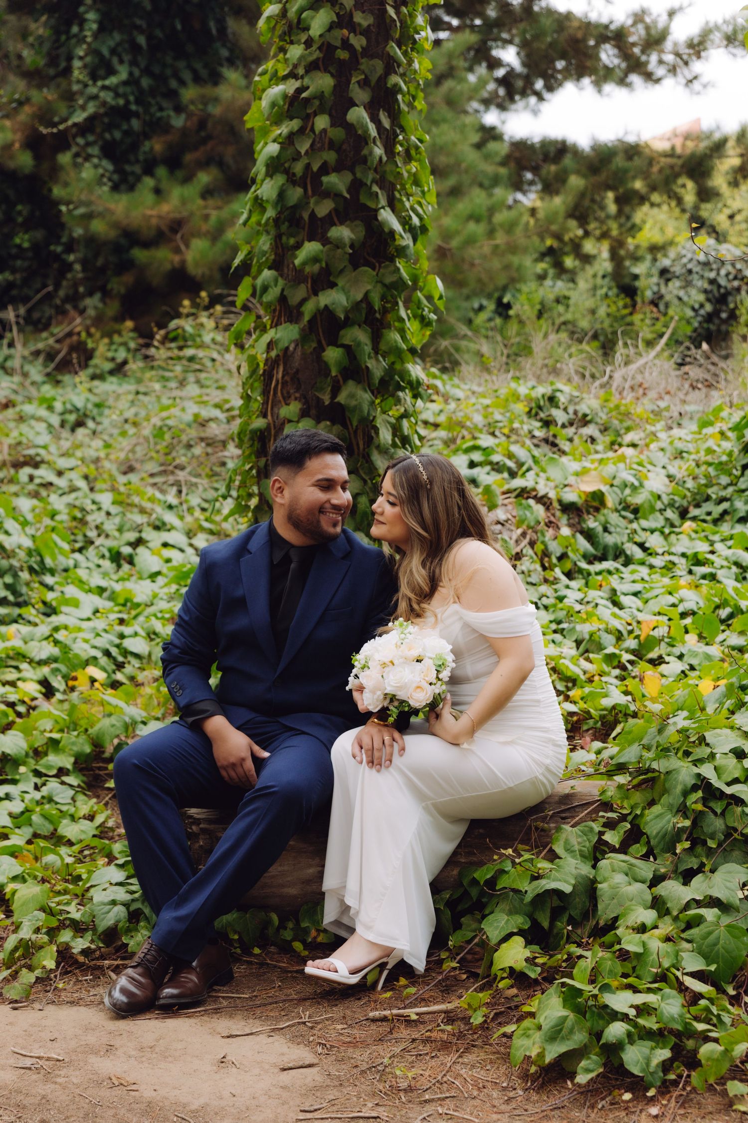 Bride and groom laugh together seated on a log bench surrounded by green ivy, bride holding white bouquet.