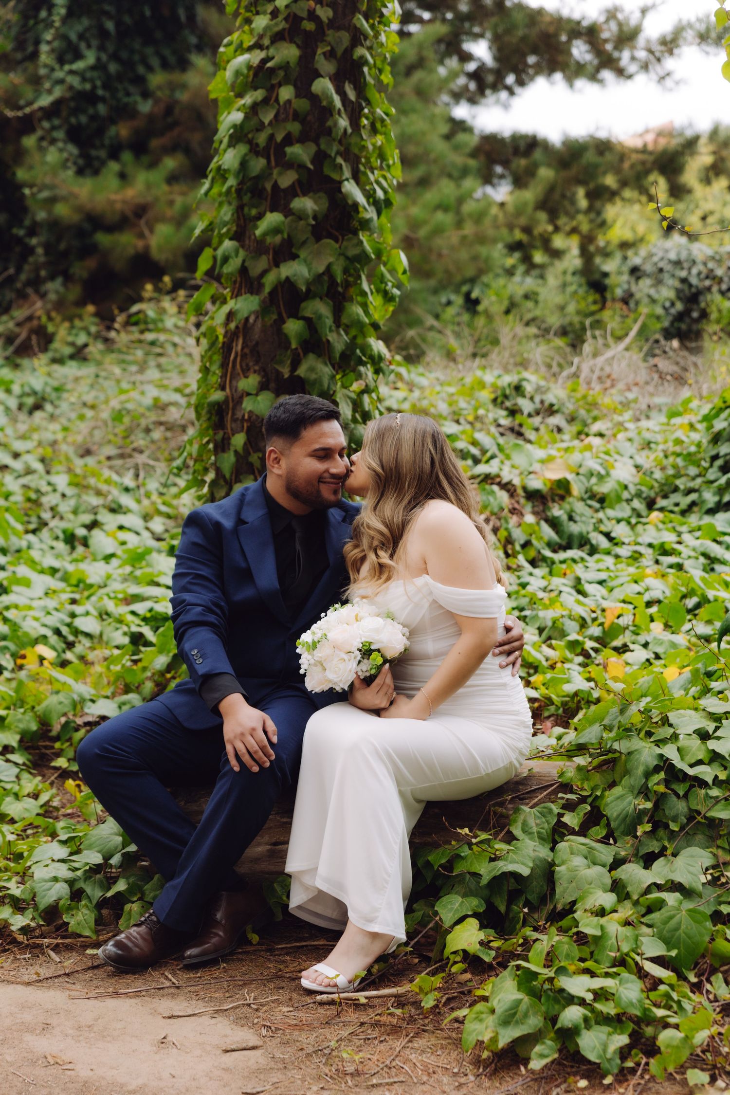 Wedding couple shares a tender kiss on a log bench beneath an ivy-covered tree in a lush garden.
