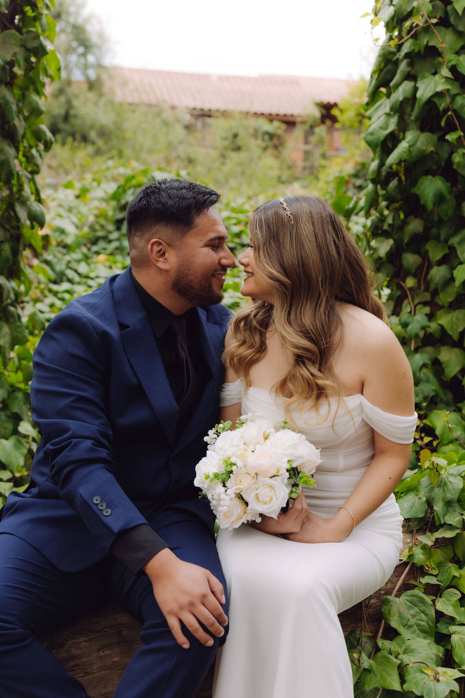 Bride and groom smile intimately at each other seated among green vines, bride holding white peony bouquet.