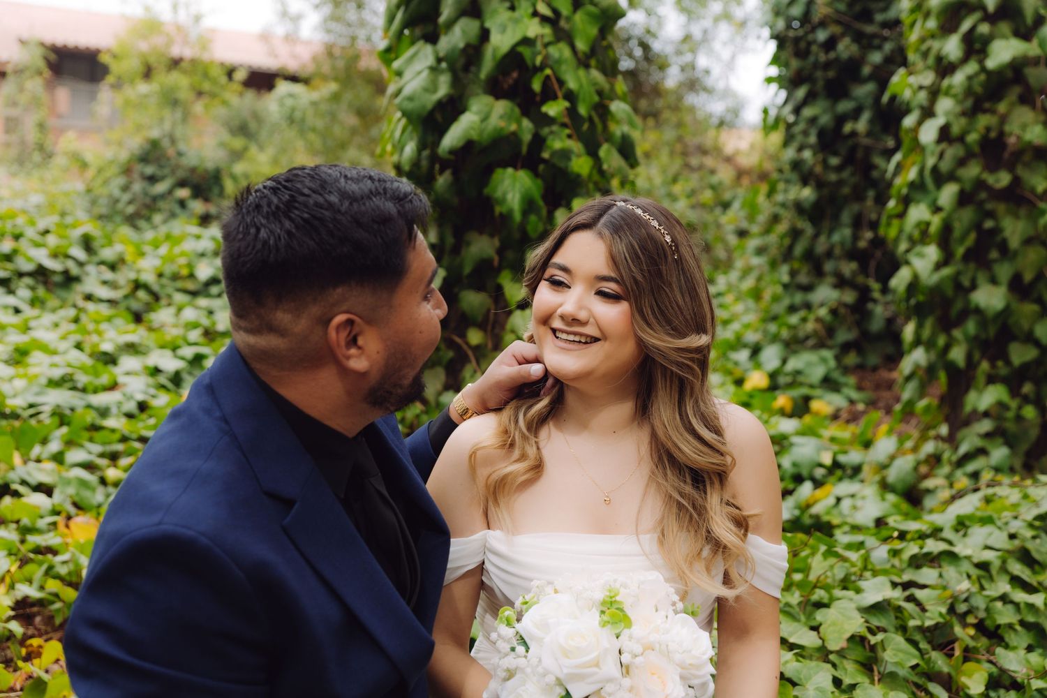 Groom tenderly touches brides face as she smiles, both surrounded by lush green garden foliage.
