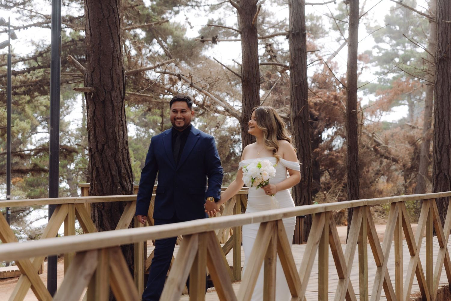 Bride and groom holding hands walking on a wooden bridge surrounded by tall pine trees.