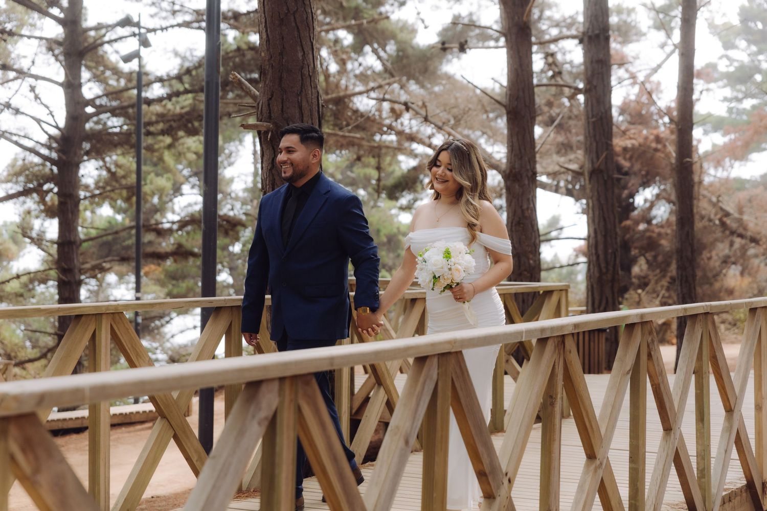A bride and groom walk hand in hand on a wooden bridge surrounded by tall pine trees.