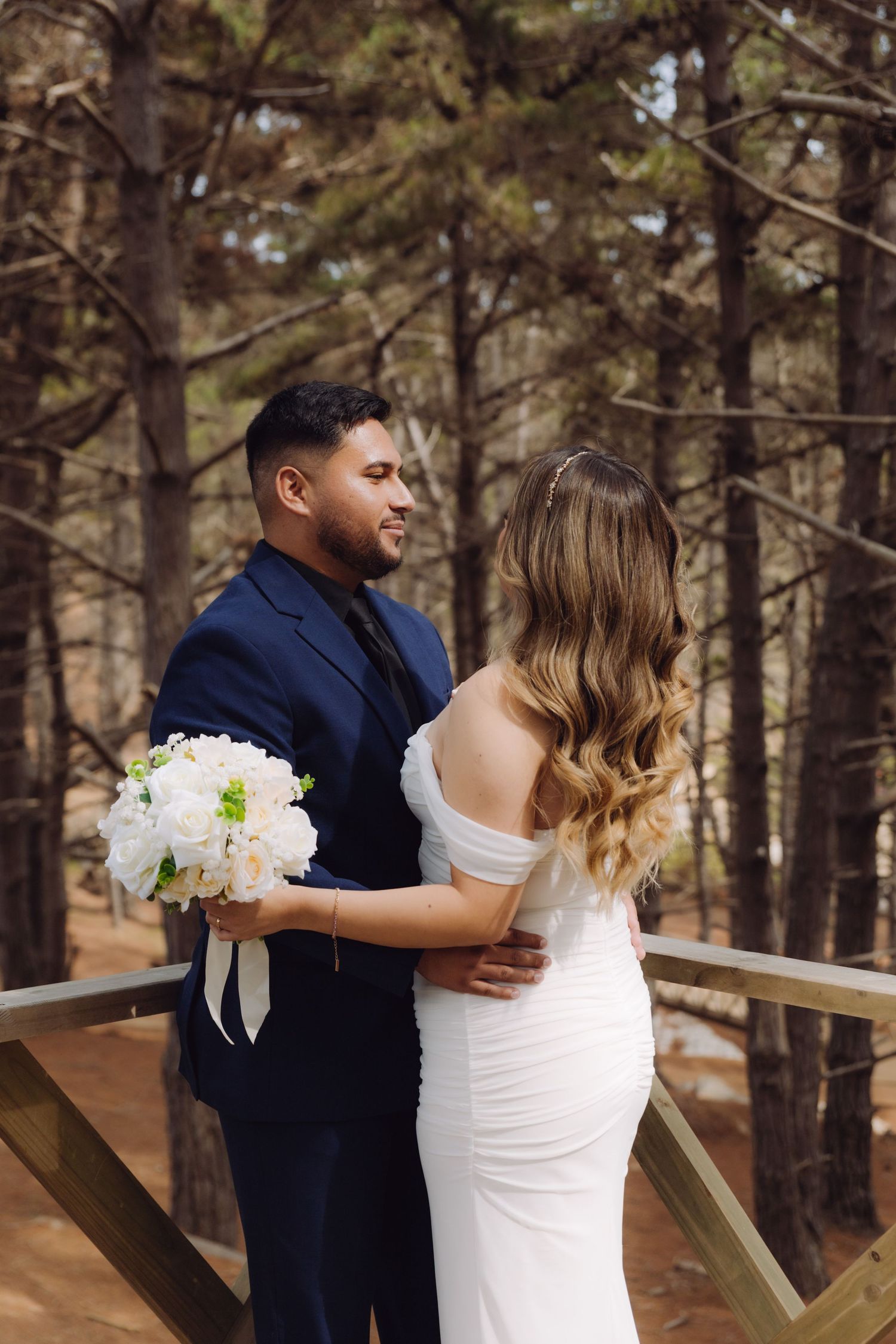 A groom in a navy suit holds a white bouquet while embracing his bride in a forest setting.