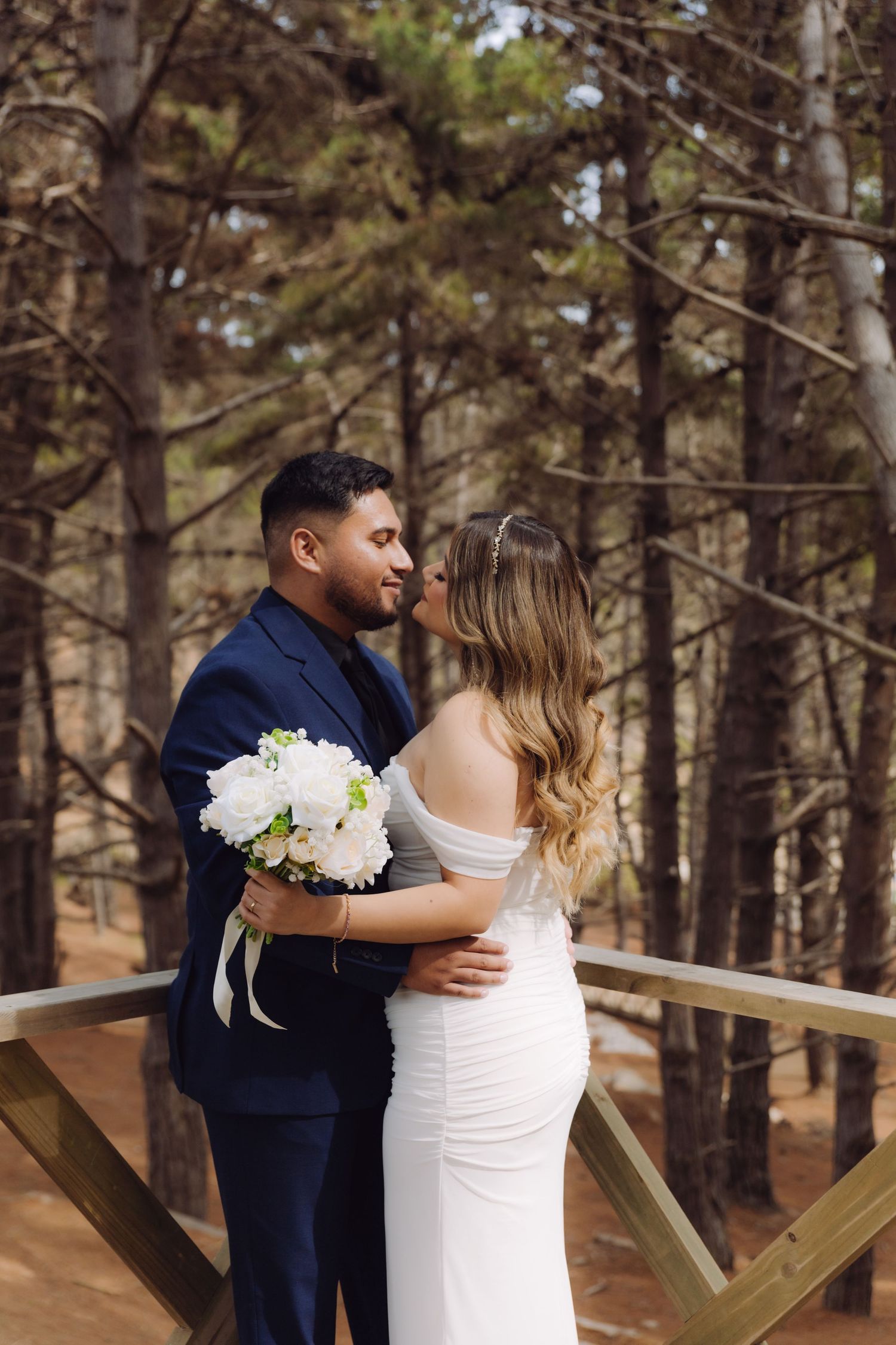 A wedding couple shares a romantic moment on a wooden deck surrounded by tall pine trees.