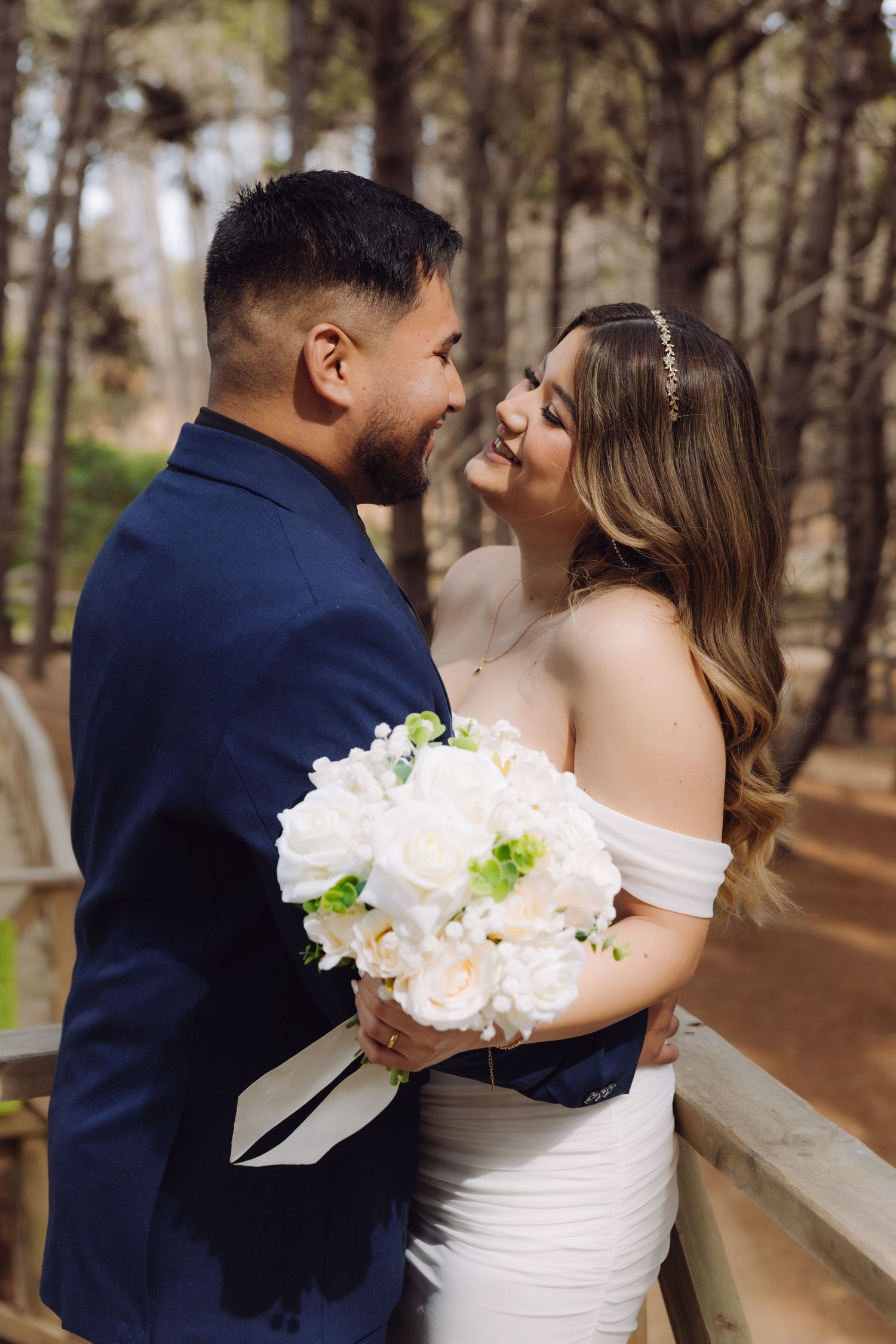 A smiling bride and groom face each other holding a white rose bouquet in a pine forest.