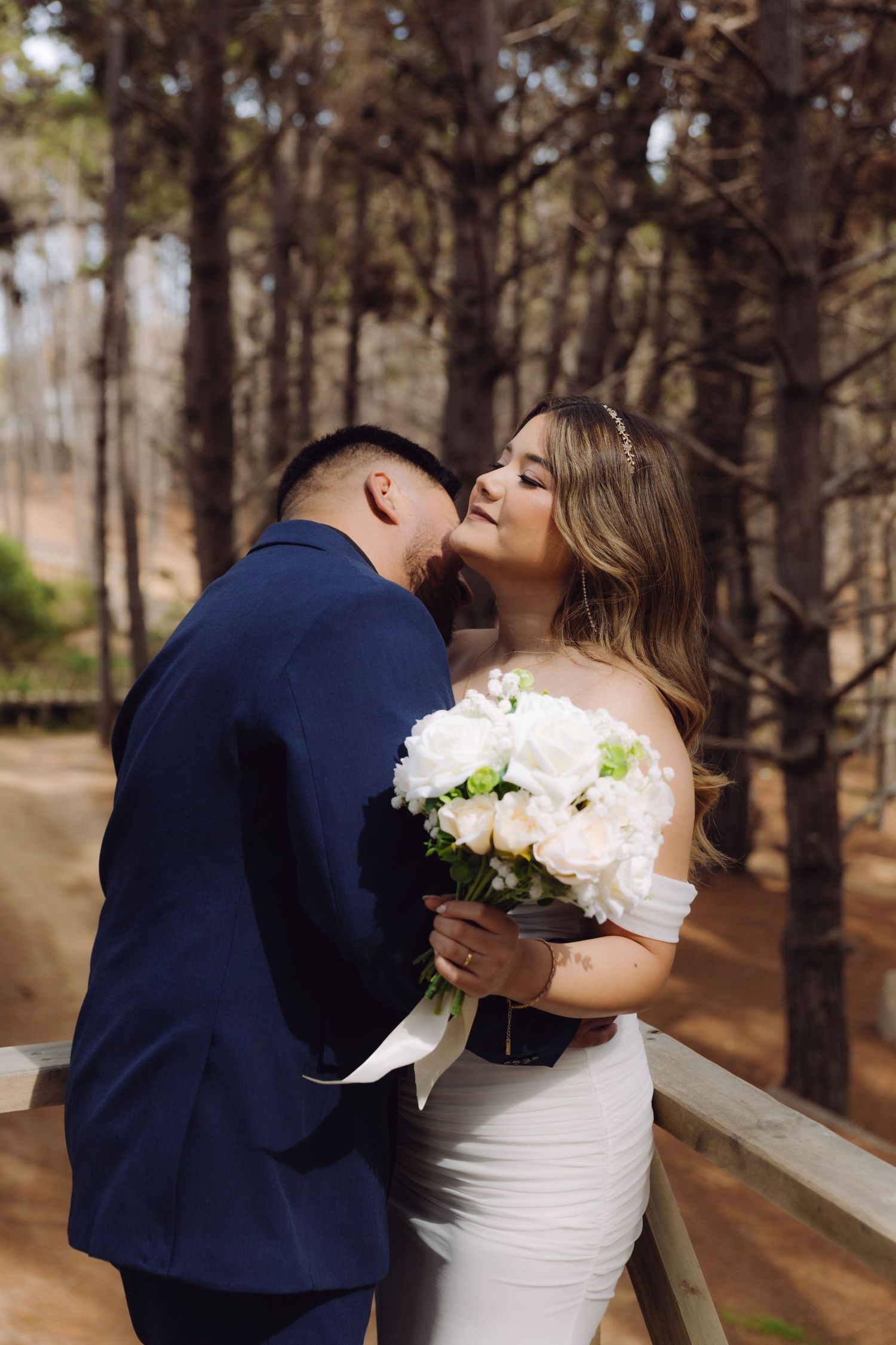 A bride and groom share a tender kiss while holding a white peony bouquet in a forest.