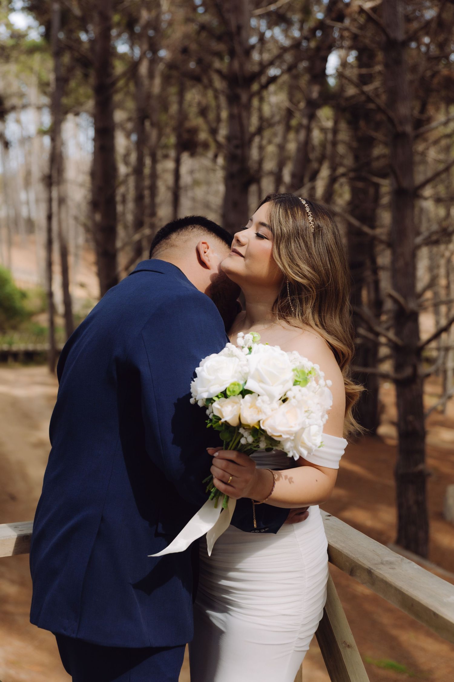 A groom kisses his bride on the cheek as she smiles holding a white bouquet in a forest.