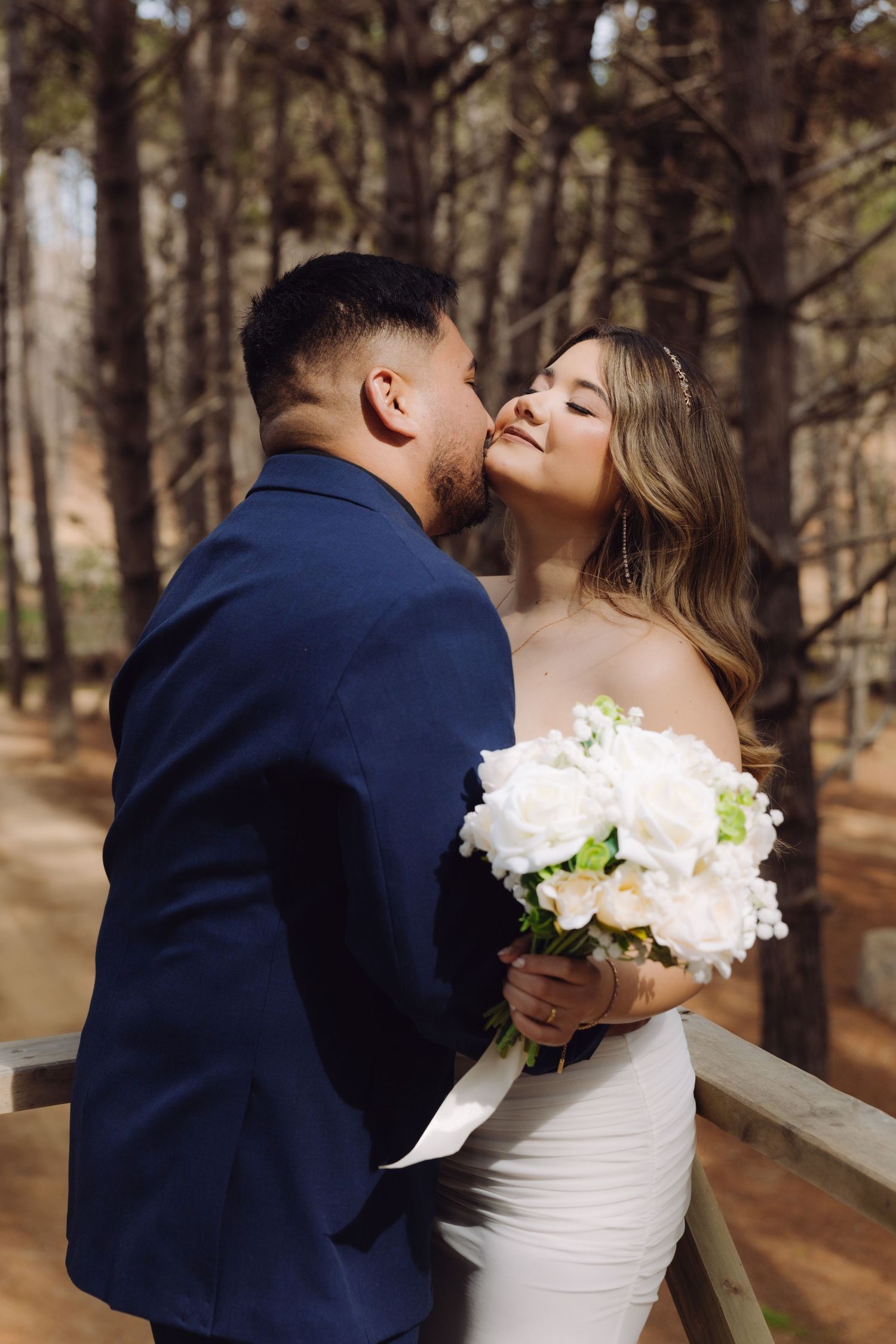 A groom in a navy suit kisses his smiling bride holding a white bouquet in a pine forest.