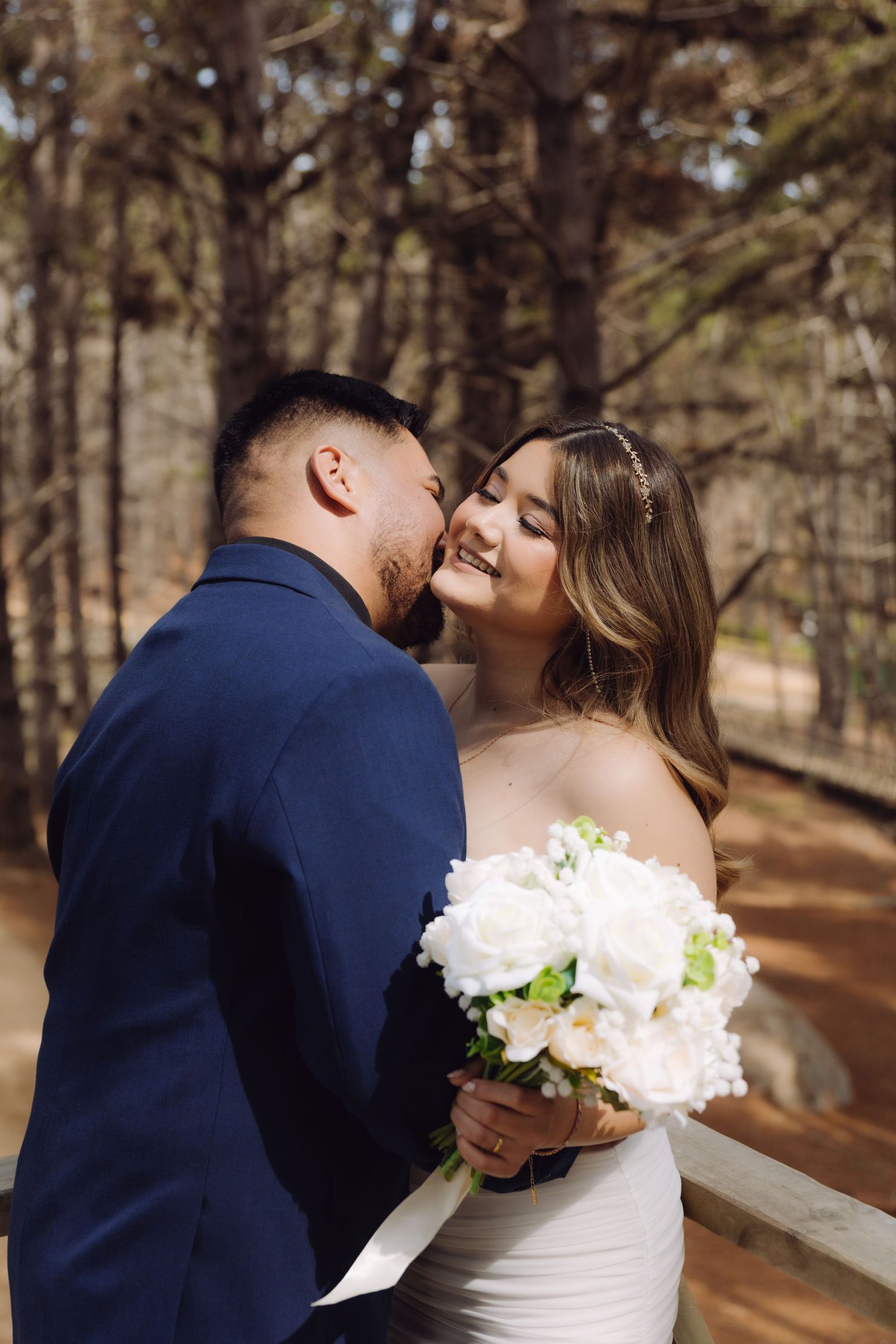 A groom whispers to his smiling bride holding a white bouquet on a sunny pine forest path.
