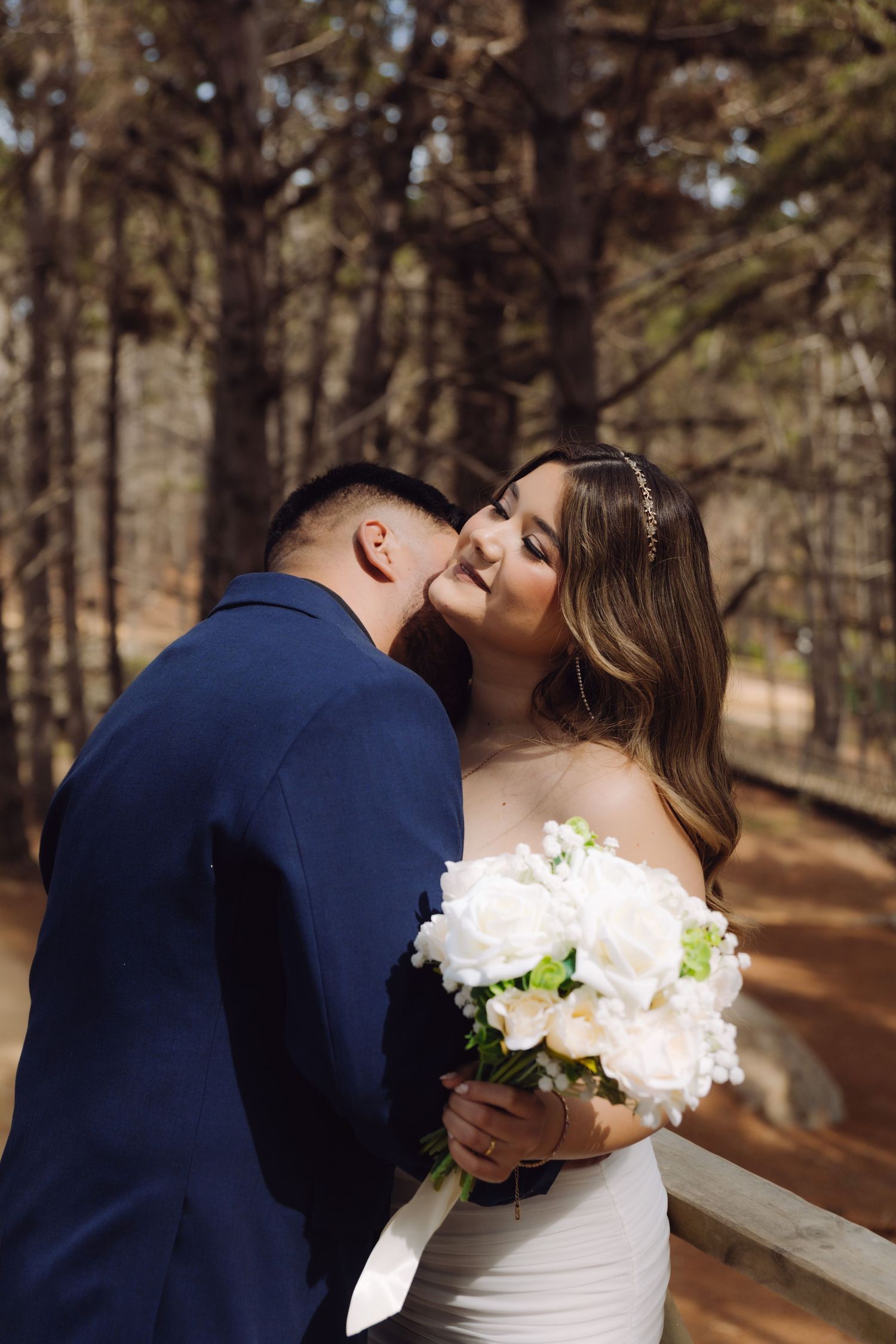 A groom kisses his bride on the cheek as she smiles holding a white bouquet in sunlit woods.