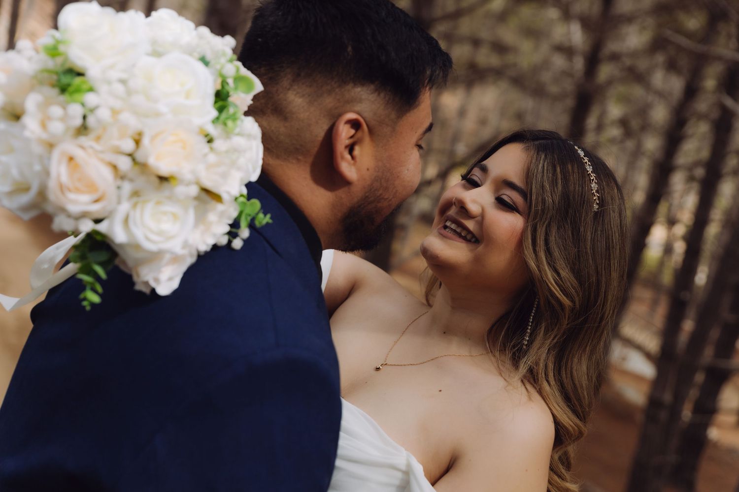 Close-up of smiling bride with floral headpiece and groom in navy suit sharing intimate forest moment.