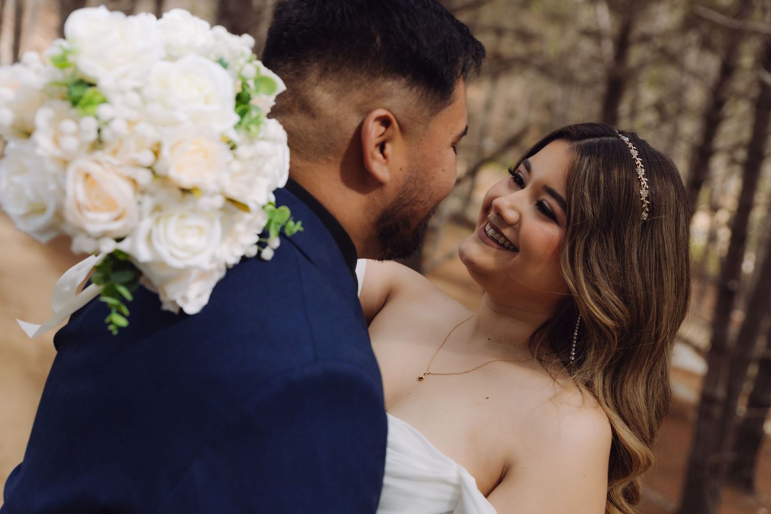 Bride smiling joyfully at groom holding white rose bouquet during outdoor forest wedding photo session.