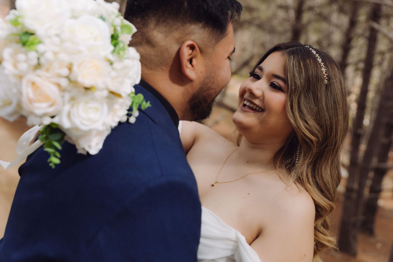 Radiant bride in white dress smiles at groom holding bouquet during romantic forest wedding shoot.