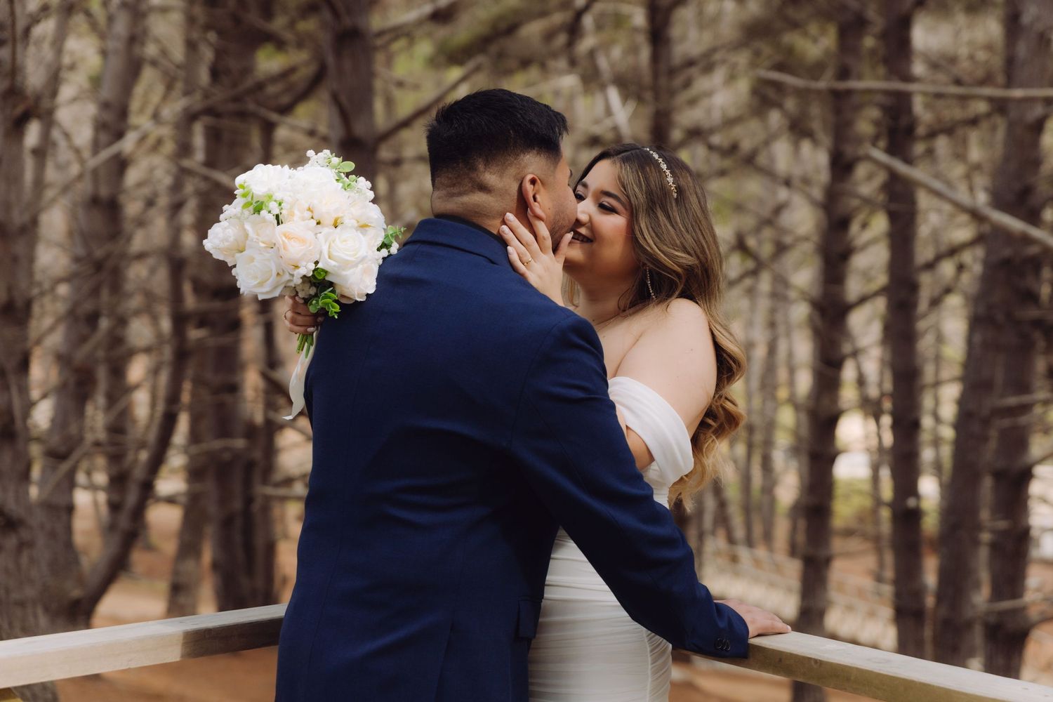 Bride and groom share a tender kiss on wooden deck surrounded by tall pine trees.