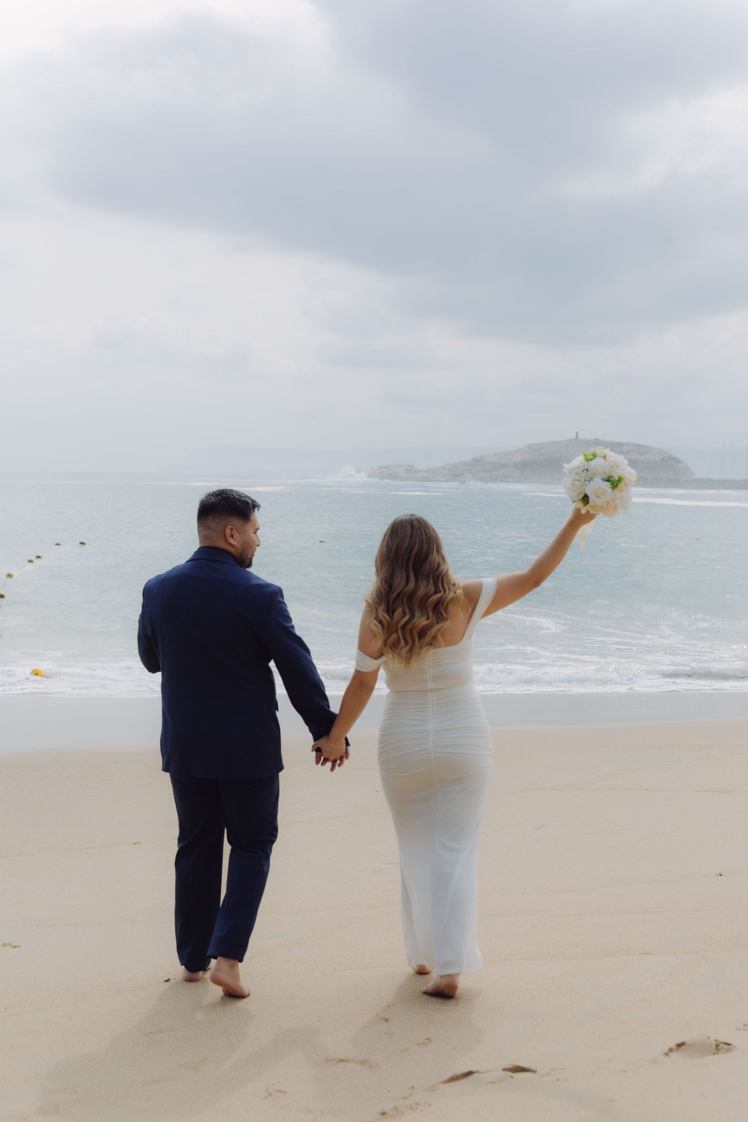Newlyweds walk barefoot on sandy beach, bride raising white bouquet toward overcast sky.