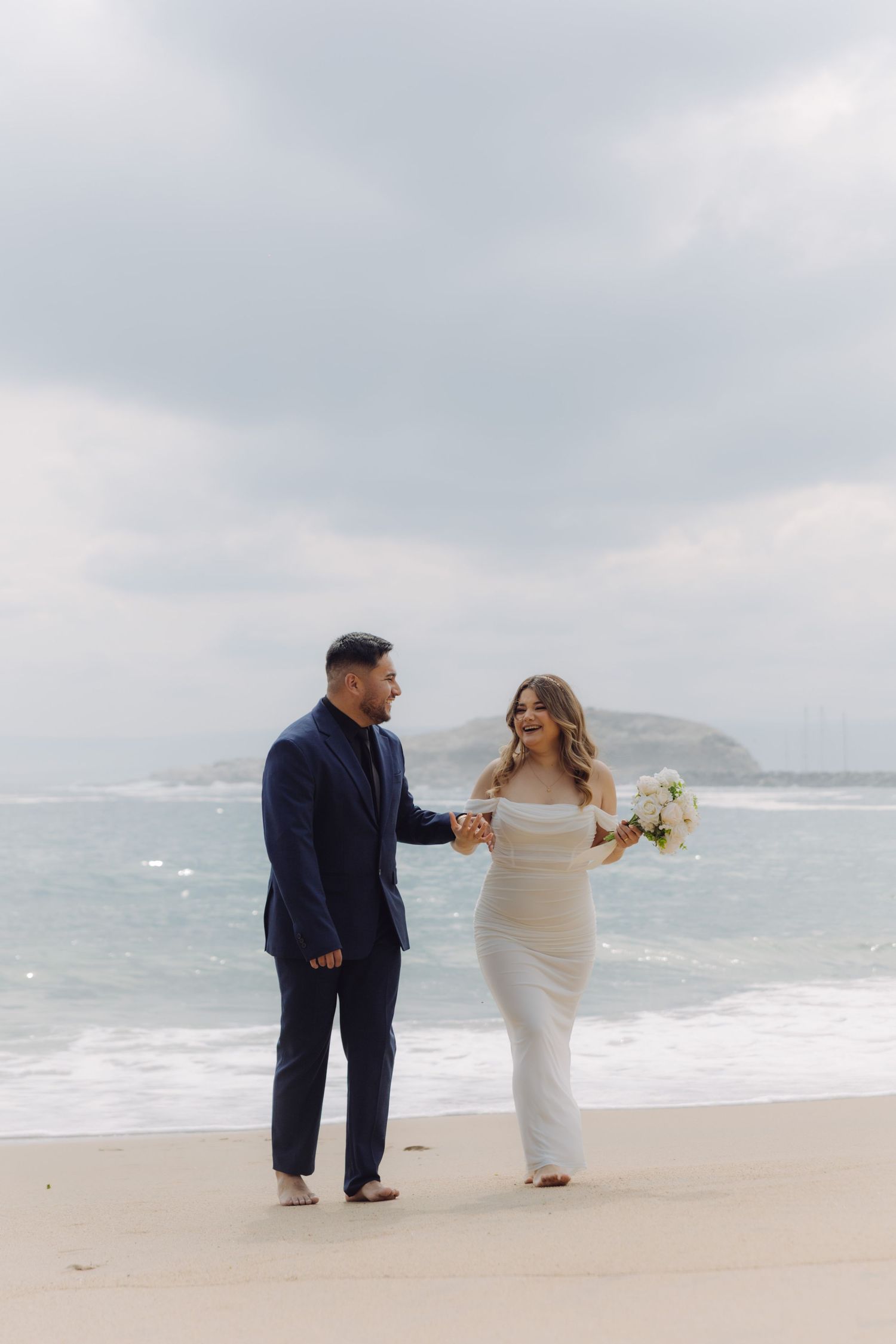 Bride and groom hold hands while walking along ocean shoreline in elegant wedding attire.