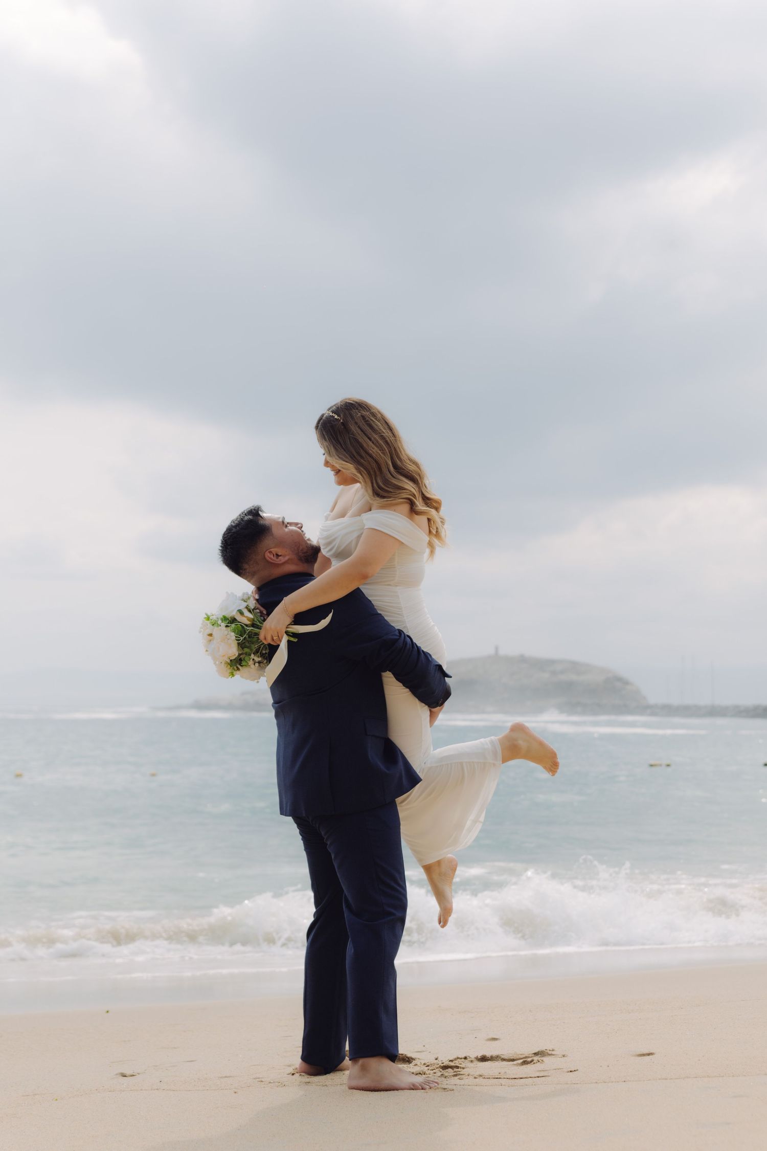 Groom lifts bride joyfully on sandy beach with ocean waves and cloudy sky behind them.
