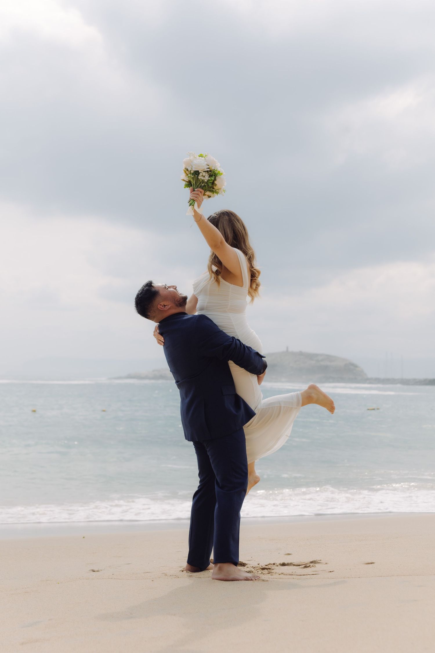 Groom lifts bride high on beach as she raises white bouquet triumphantly toward the sky.