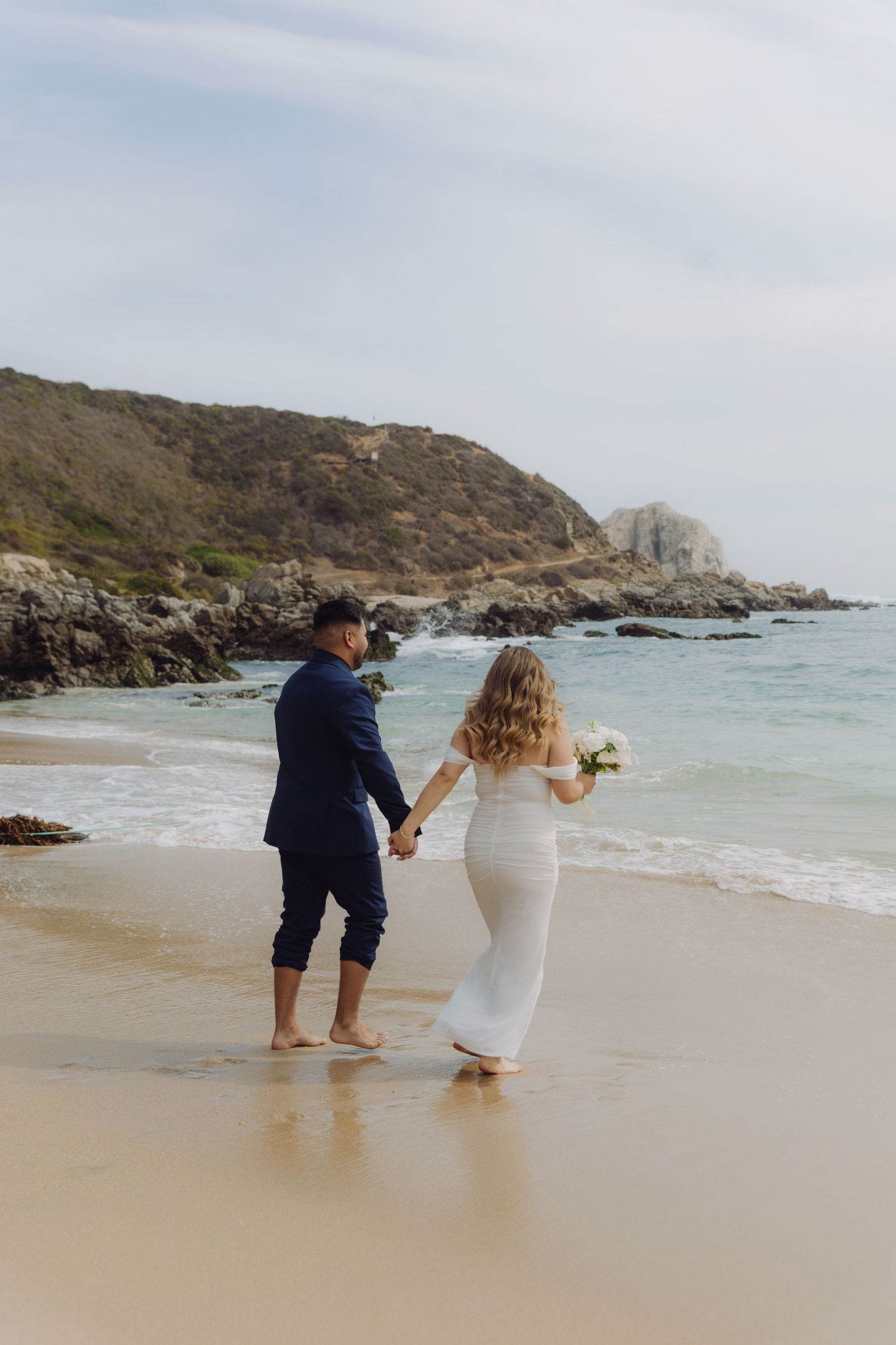 Bride and groom walking hand in hand barefoot on a sandy beach with rocky cliffs and turquoise ocean.