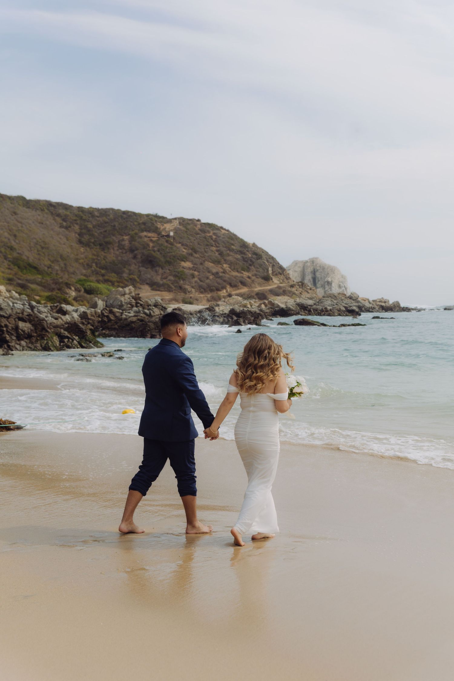 Newlywed couple strolling along the shoreline on a beach, bride holding white bouquet in white gown.