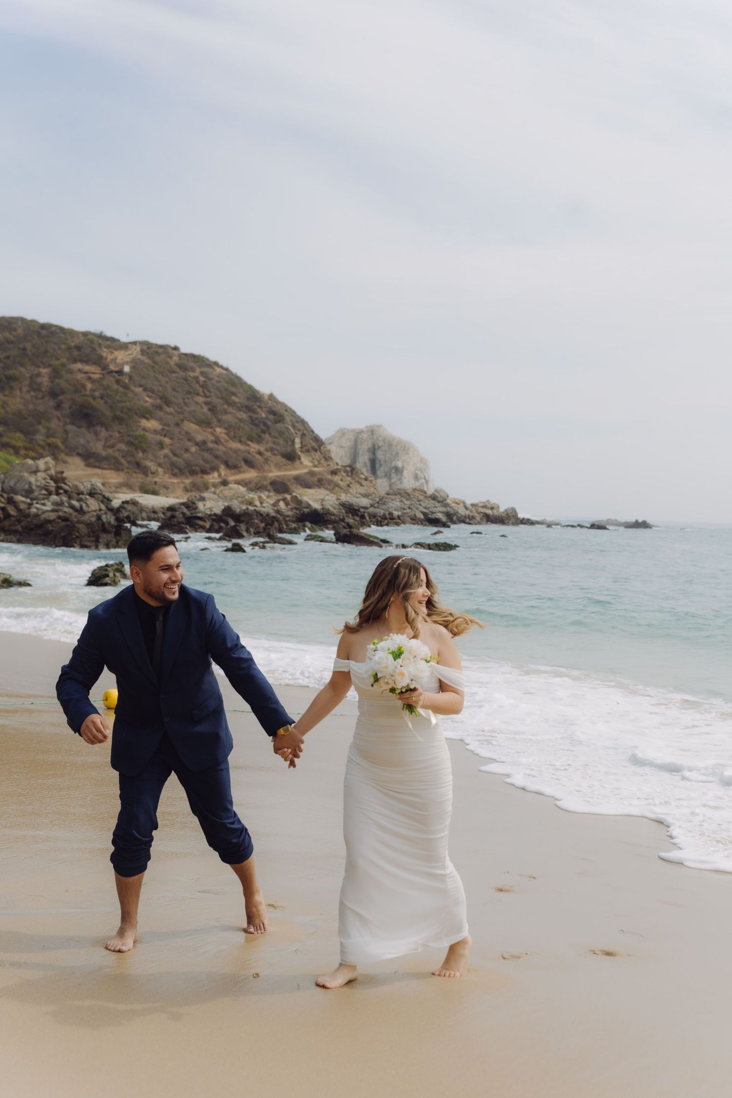 Happy bride and groom walking barefoot on a scenic beach, bride carrying white floral bouquet.