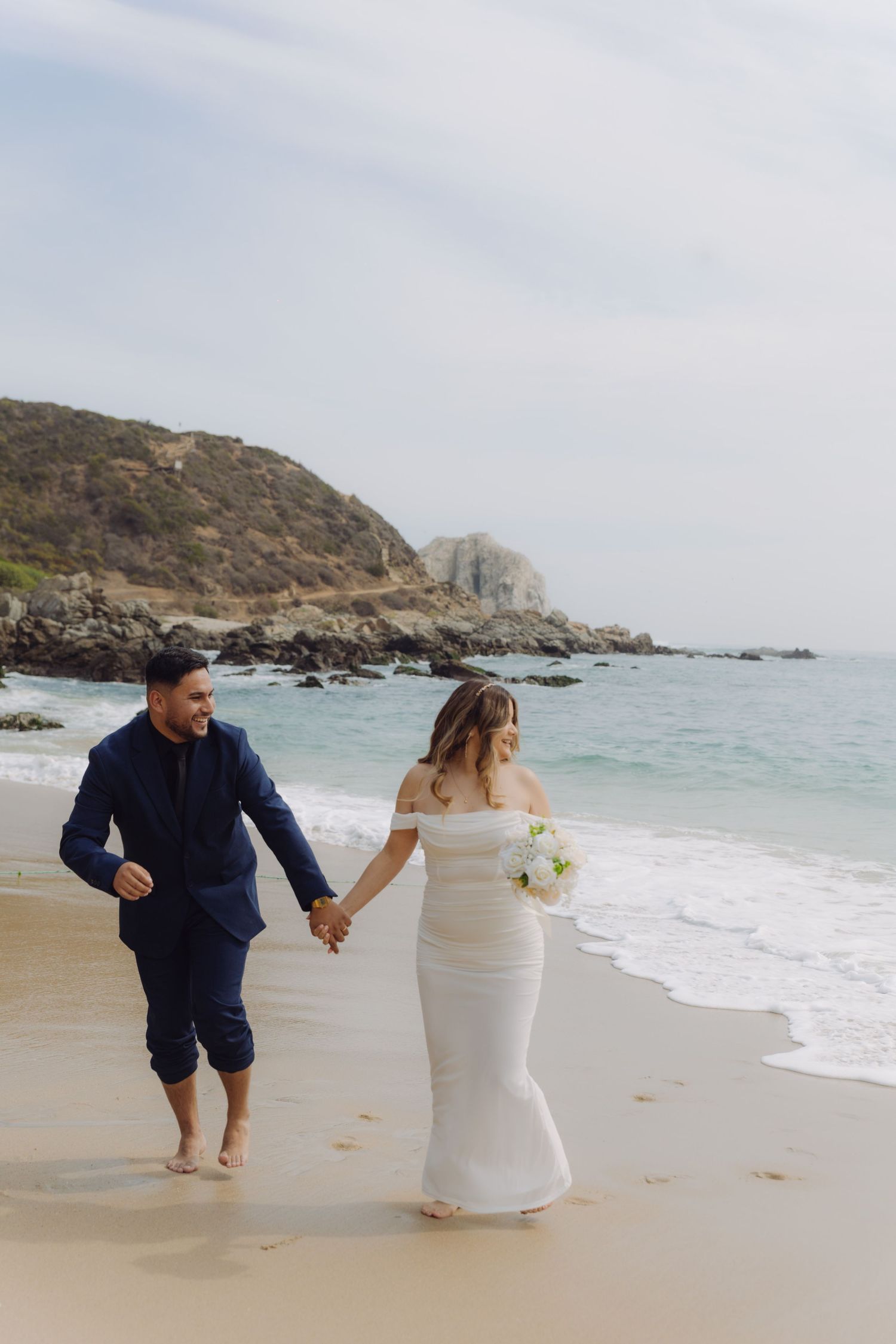 Couple in wedding attire holding hands and walking along sandy beach with rocky coastline backdrop.
