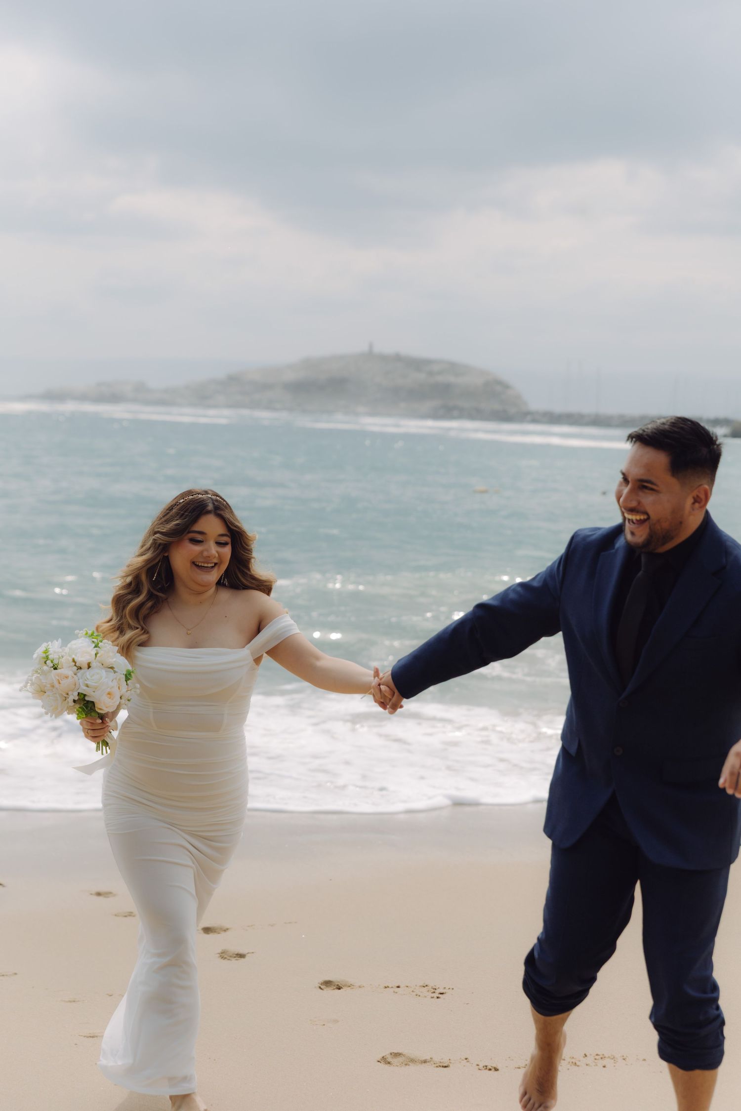 Laughing newlywed couple running hand in hand along sandy beach with ocean waves behind them.