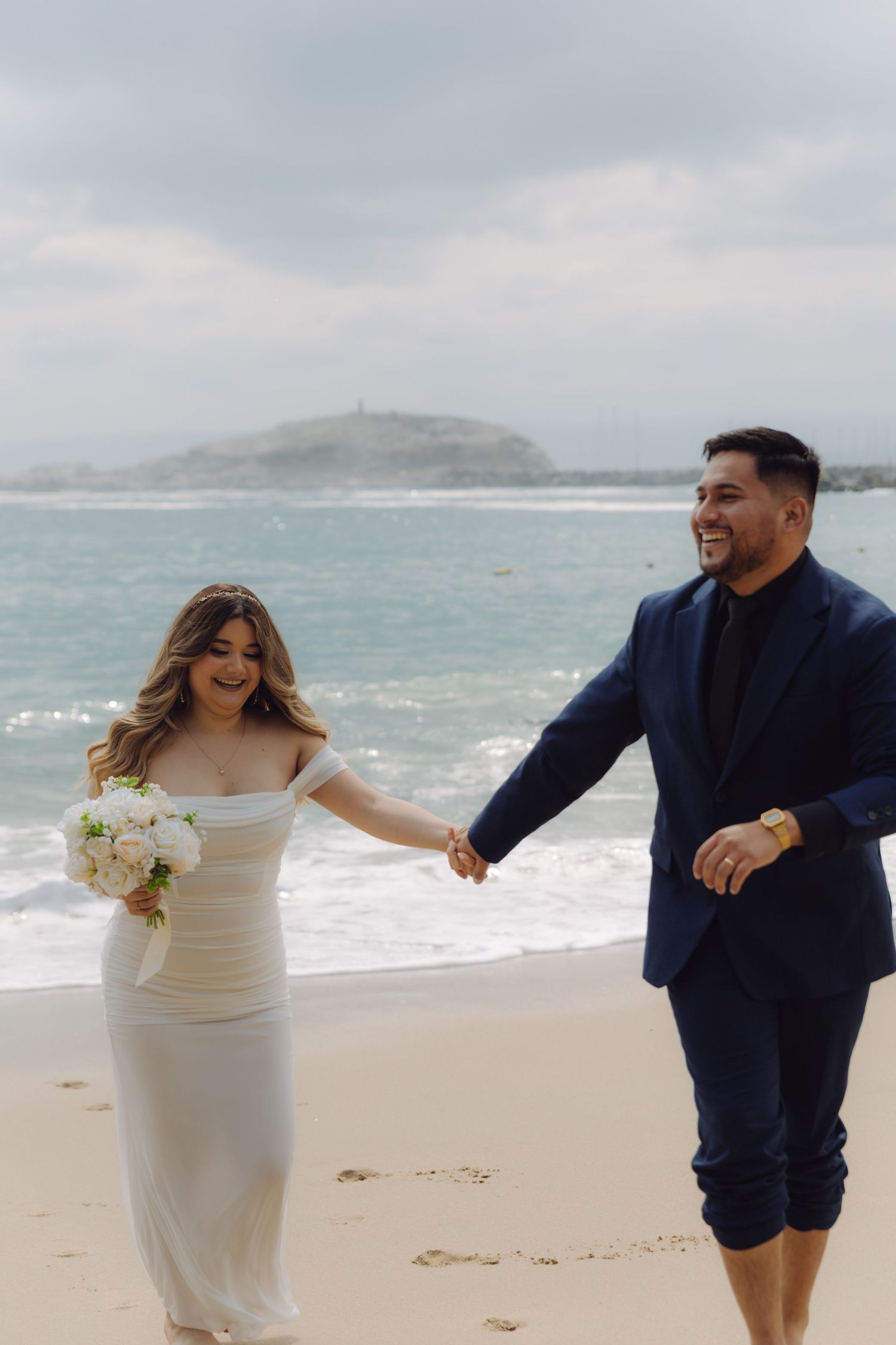 Smiling bride and groom holding hands on a beach, bride in white off-shoulder gown with bouquet.