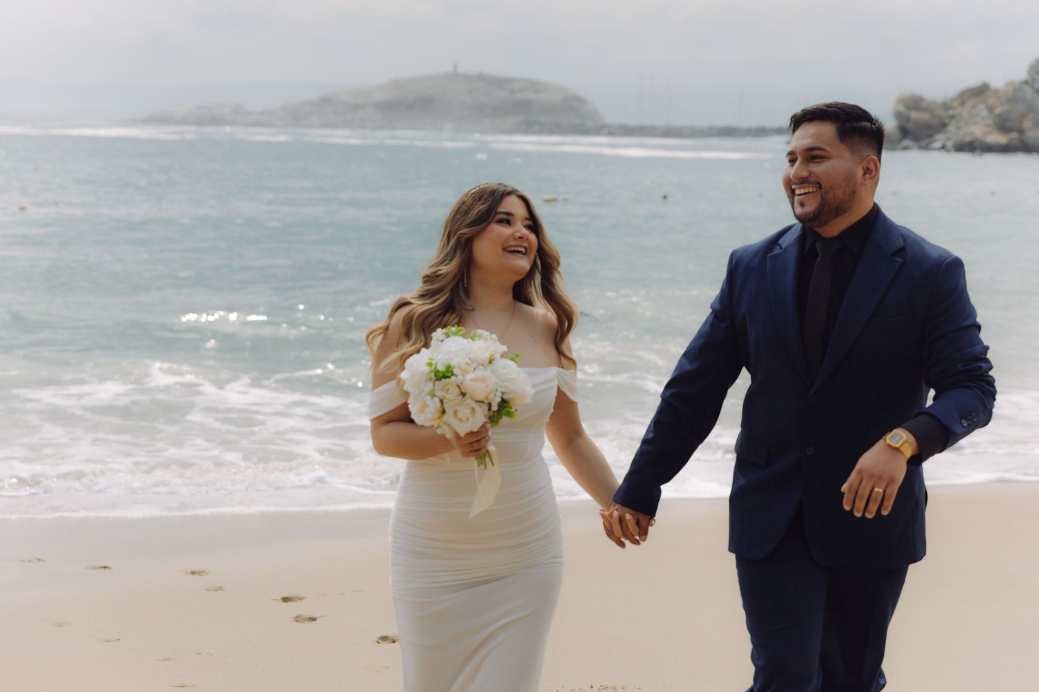 Joyful bride and groom holding hands and walking on a sunny beach, bride carrying white bouquet.