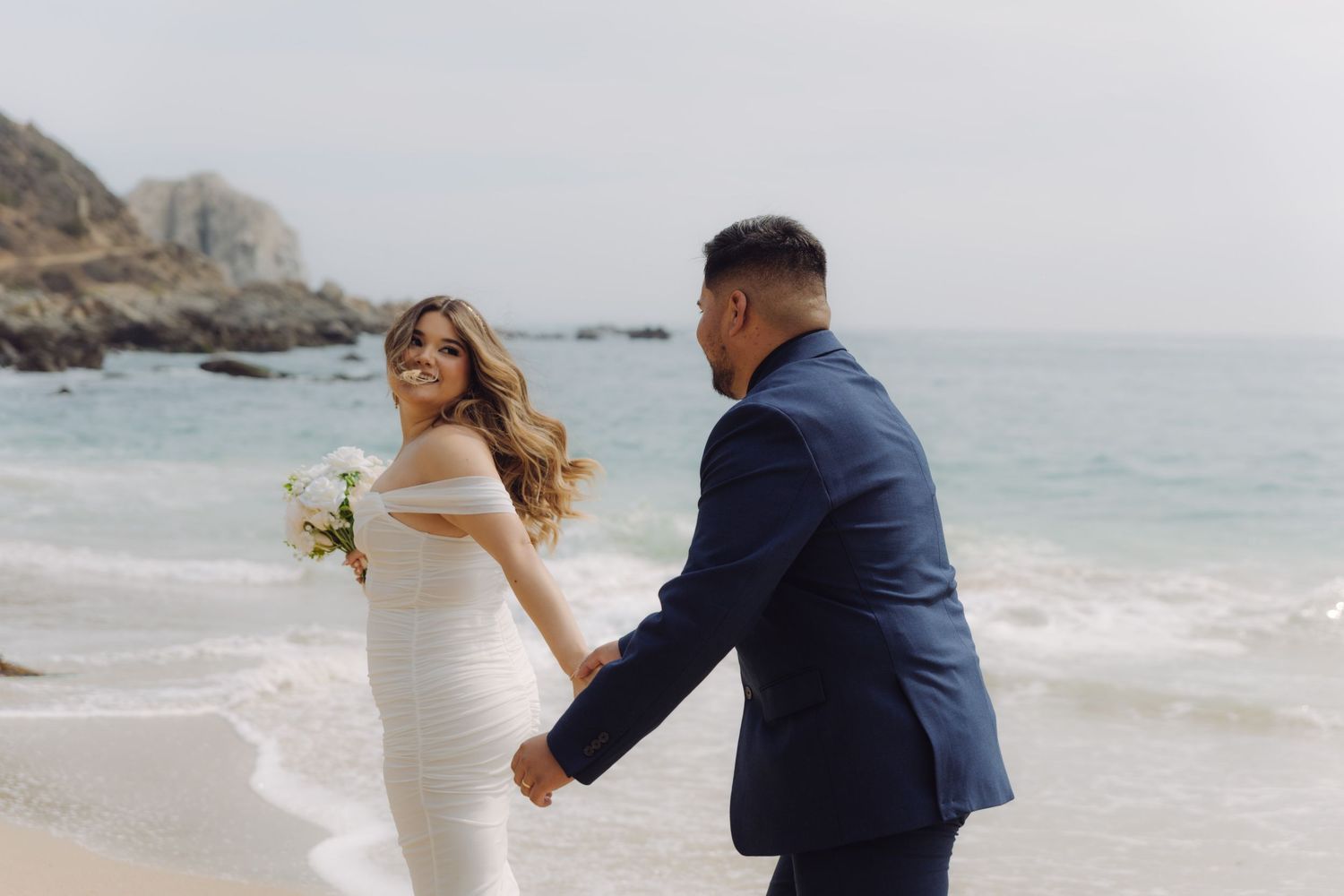 Bride in white gown looking back at groom while holding hands on a scenic rocky beach.