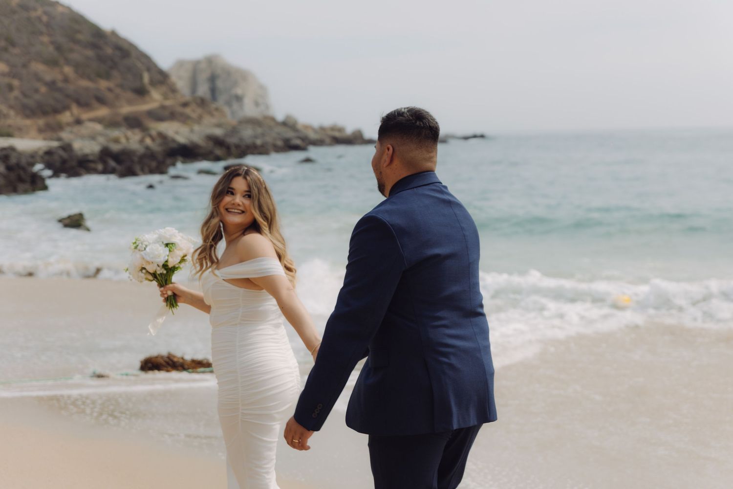 Bride glancing over shoulder at groom while holding hands on a beautiful beach with blue water.